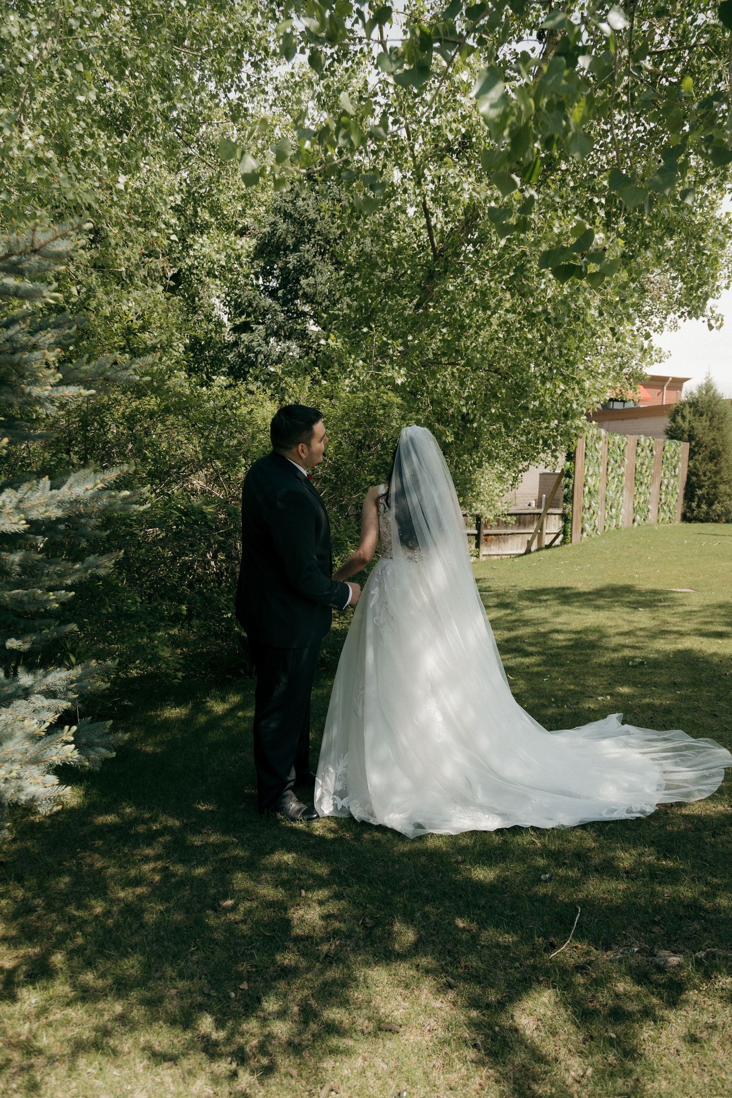 Bride and groom sharing a tender moment under green trees in a sunlit garden setting.