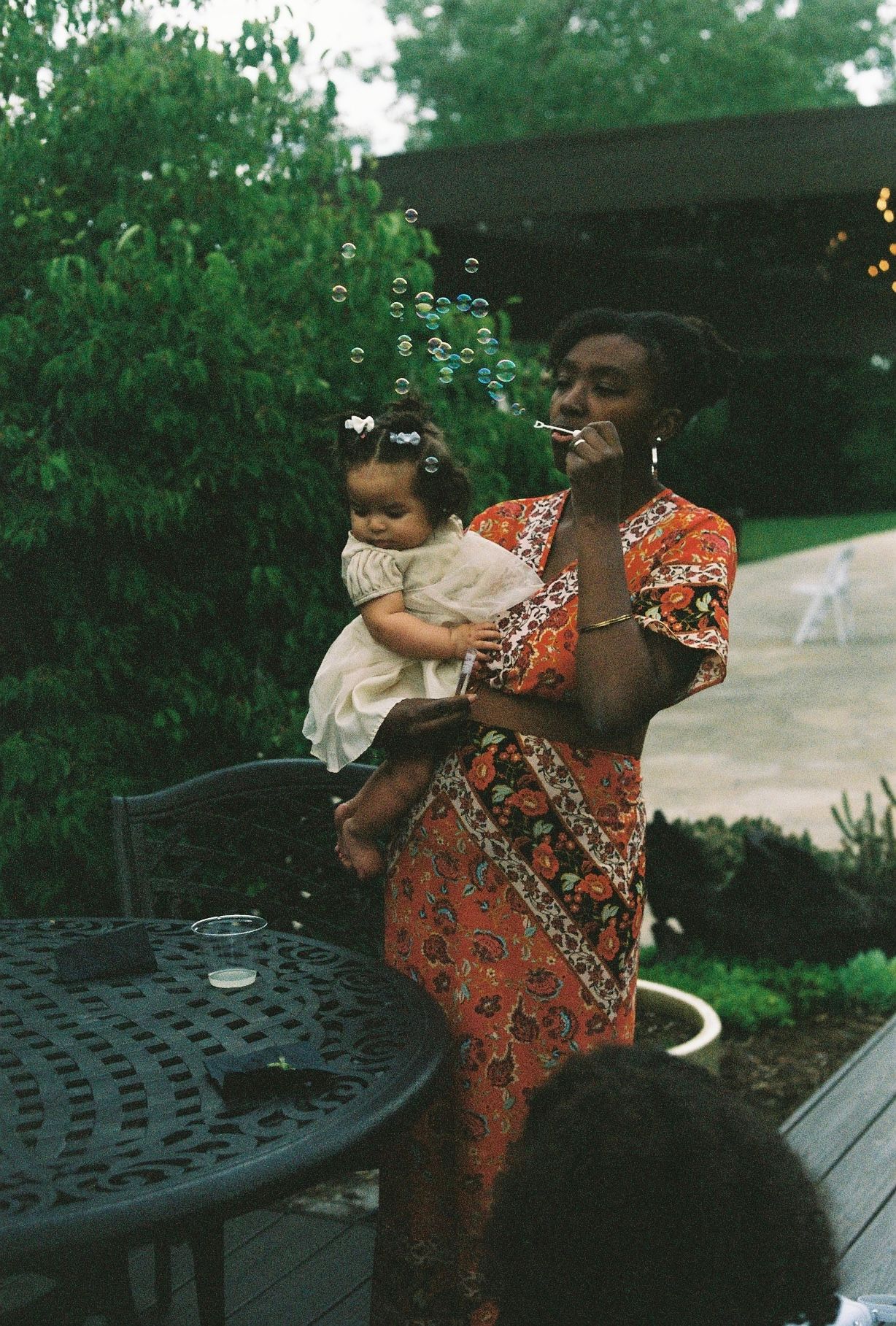 Woman in floral dress blows bubbles while holding a baby at an outdoor wedding.
