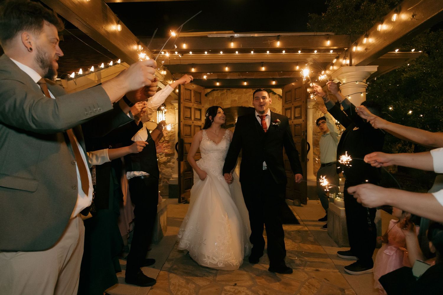 Bride and groom exit through sparkler tunnel surrounded by cheering guests at night wedding.
