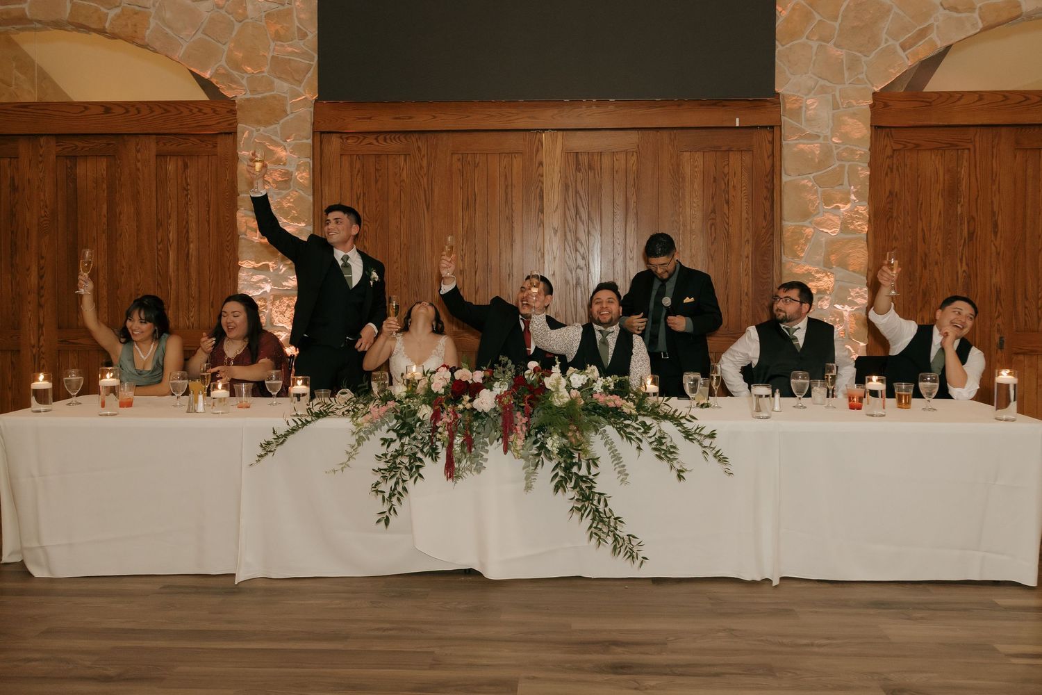 Wedding party raises hands in celebration at decorated head table with lush greenery centerpiece.