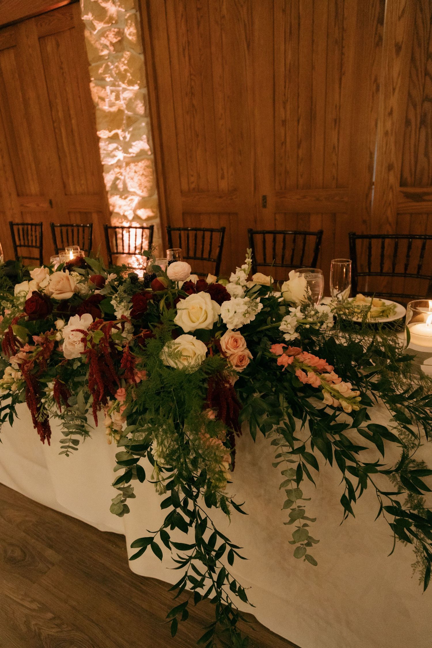 Lush wedding head table floral arrangement with burgundy, blush, and white blooms cascading over white linen.