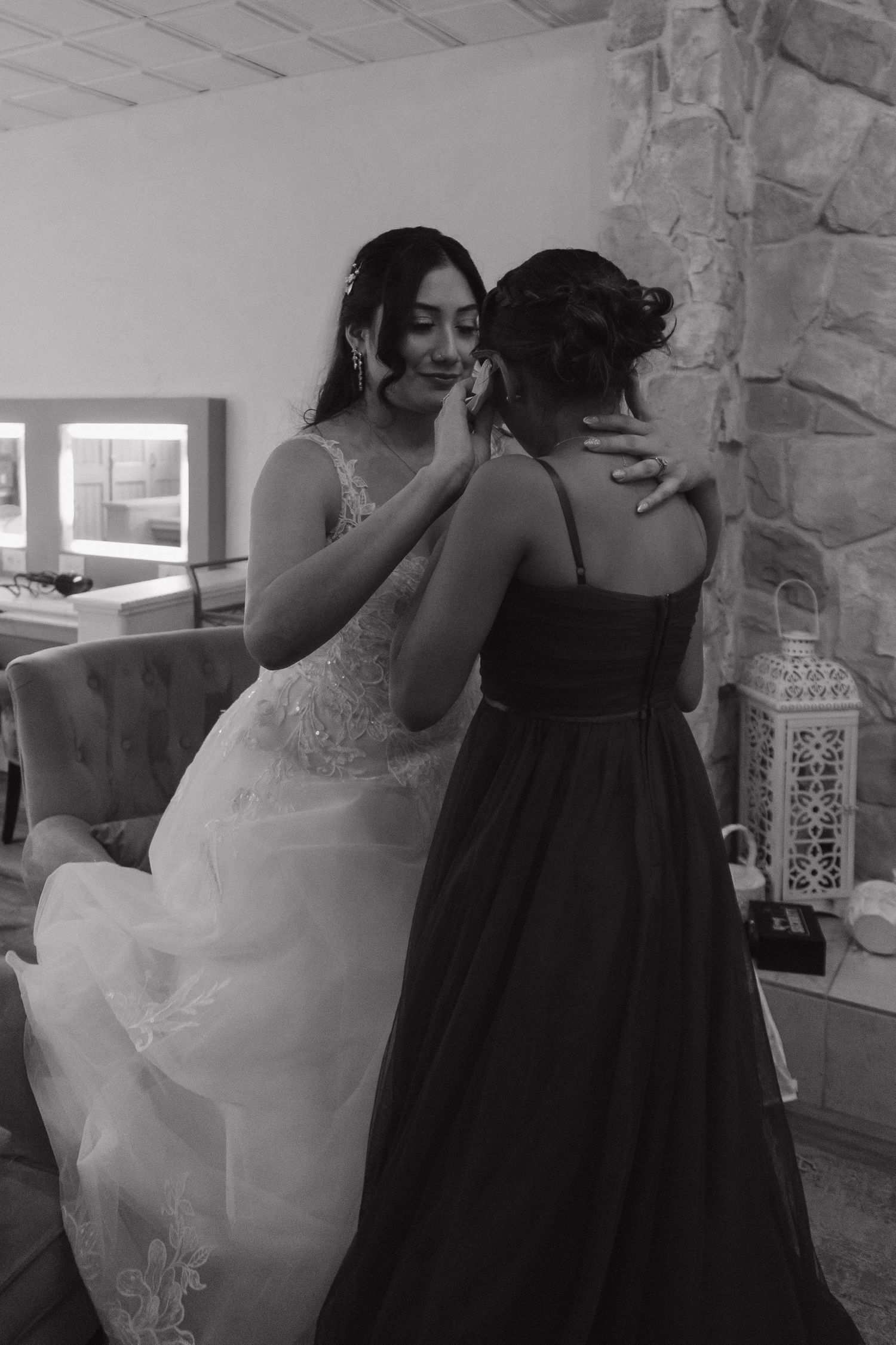 Black and white photo of bride in lace gown sharing tender moment with bridesmaid in dark dress indoors.