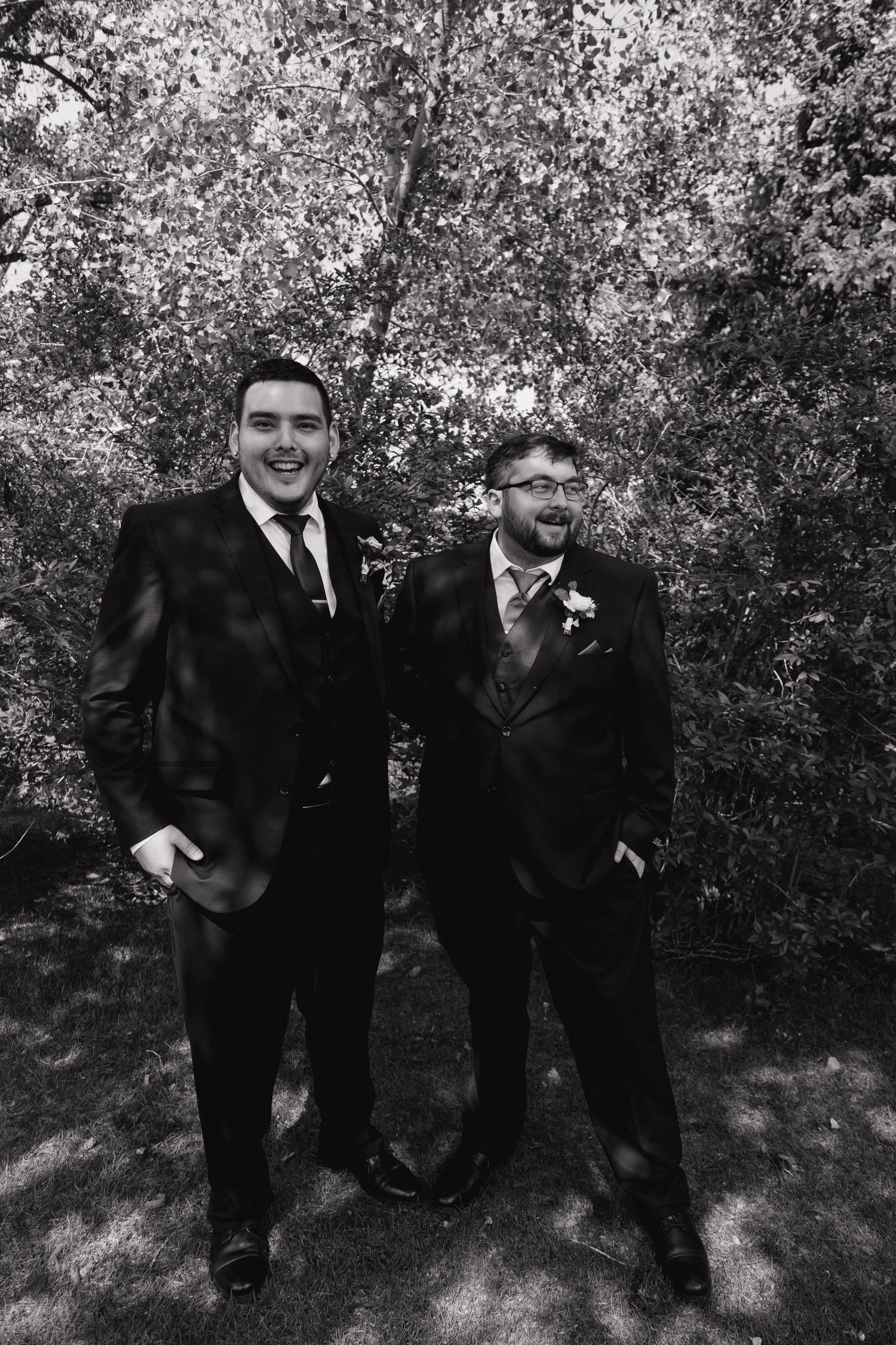 Two smiling groomsmen in black suits share a joyful moment in black and white photo.