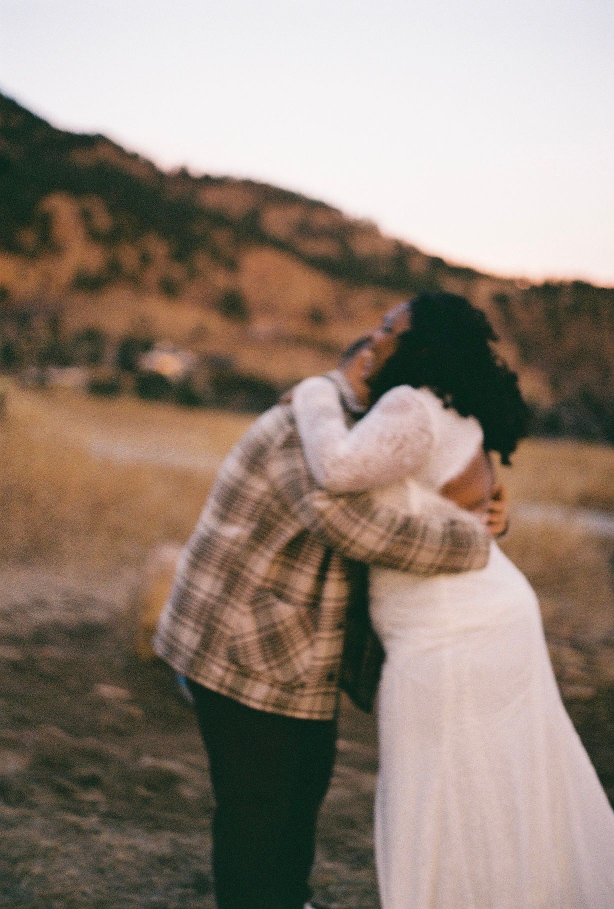 A couple in plaid and white dress embrace during sunset in a rustic outdoor setting.