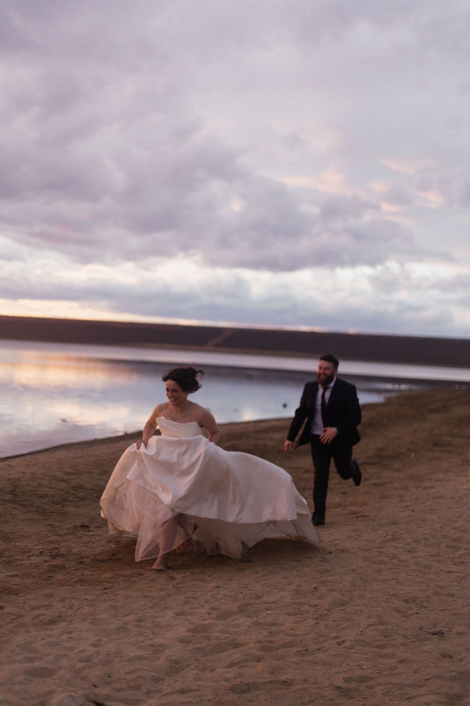 A couple runs playfully along a beach at sunset with flowing dress and formal wear.