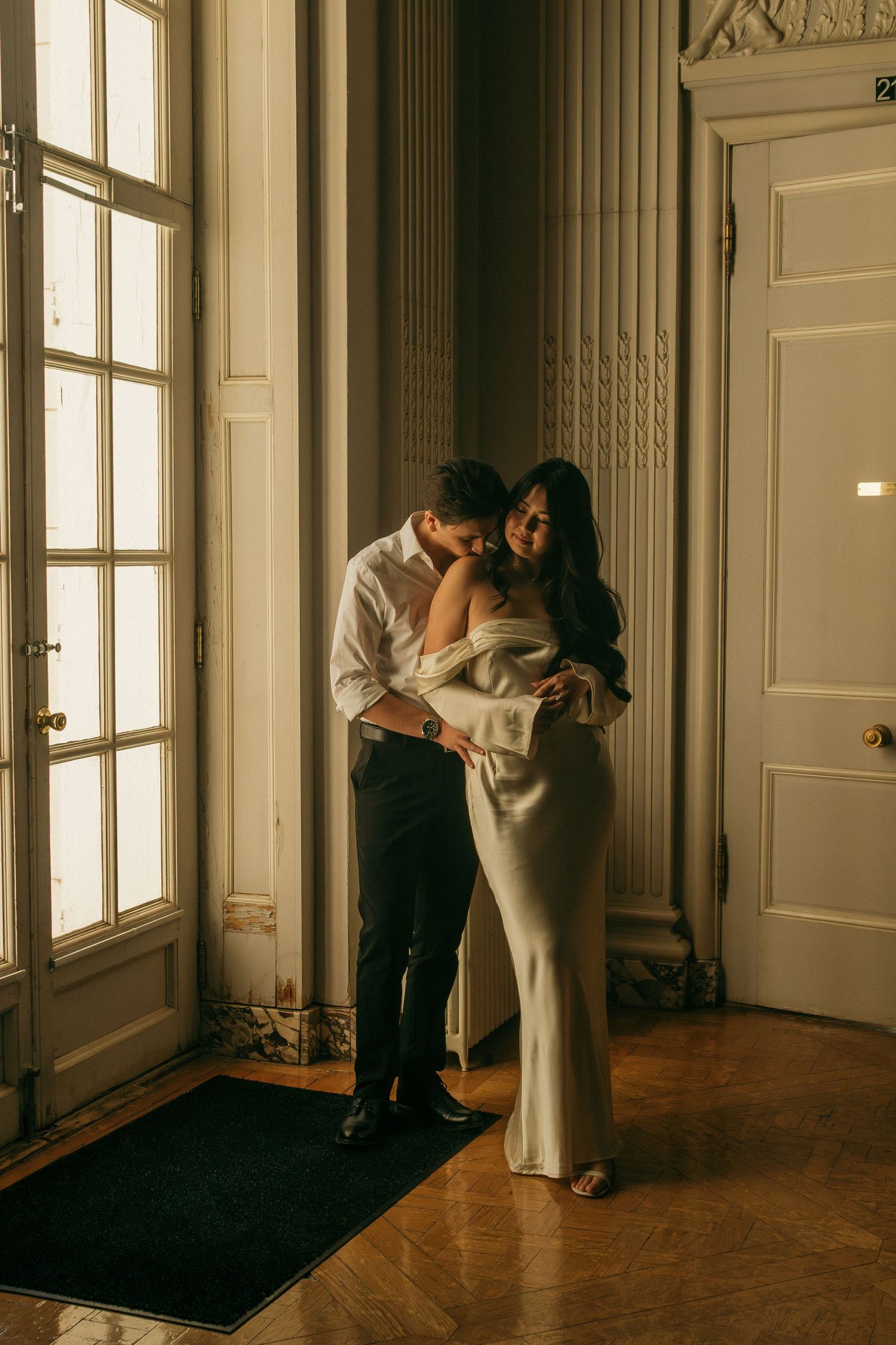 A tender moment captured in a sunlit room with hardwood floors and tall windows.