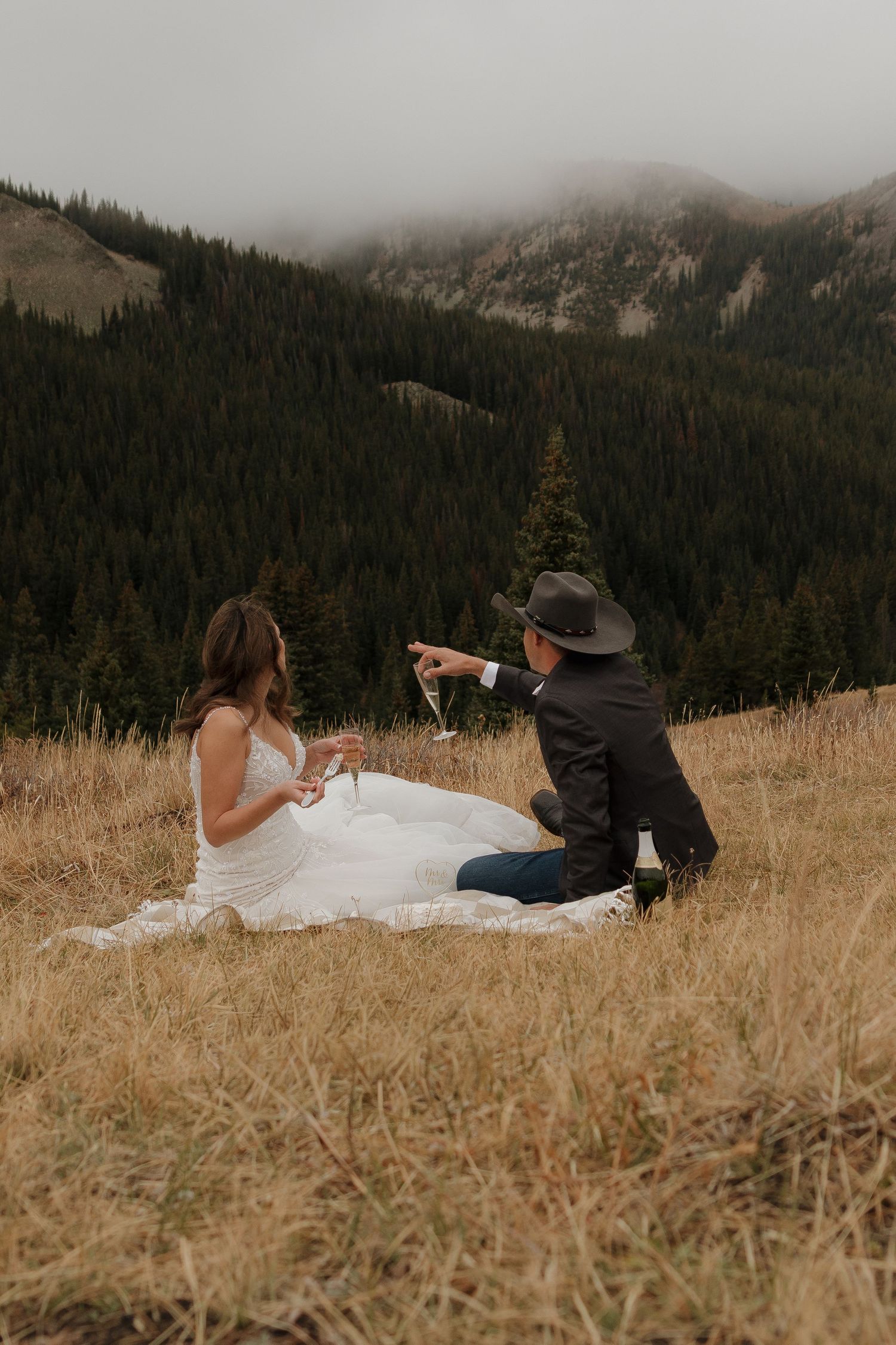 Couple enjoys champagne while sitting in a mountain meadow with dramatic landscape views.
