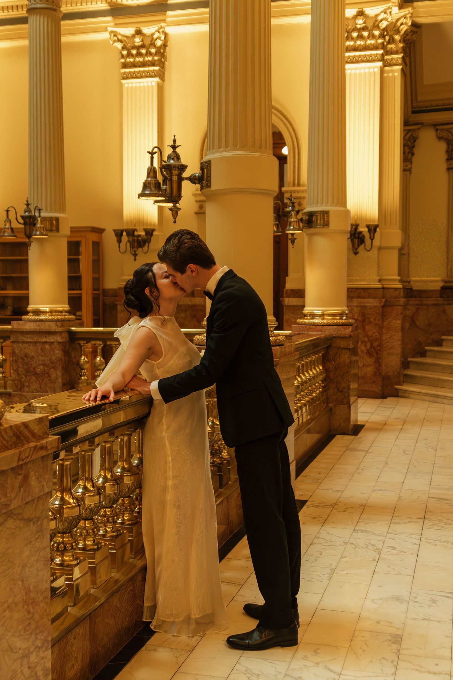 Couple in formal attire sharing an intimate moment along a marble railing in a historic building.