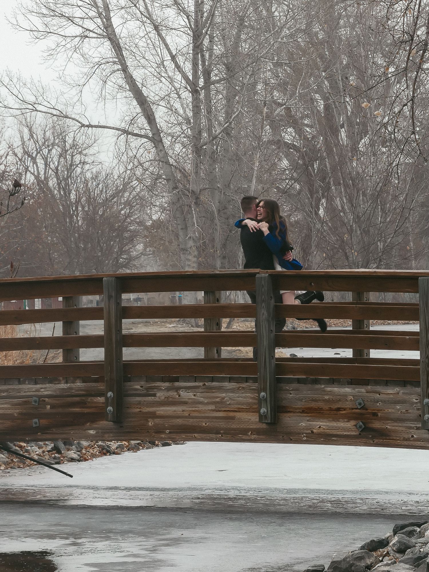 A romantic embrace on a wooden bridge during winter with bare trees in background.