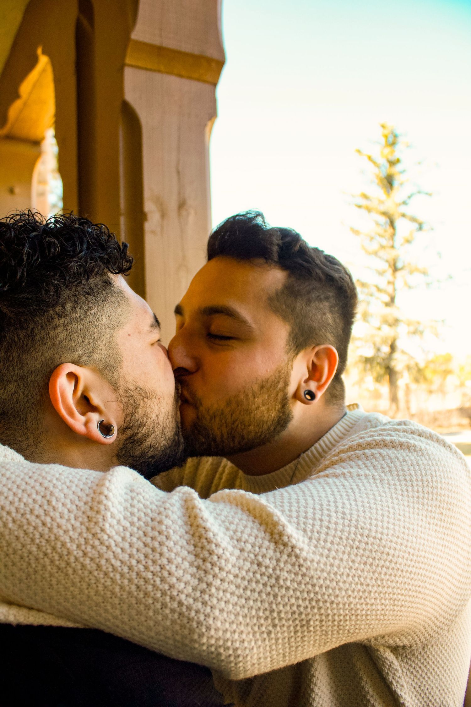 Two people share a romantic kiss outdoors with sunlit architecture in background.