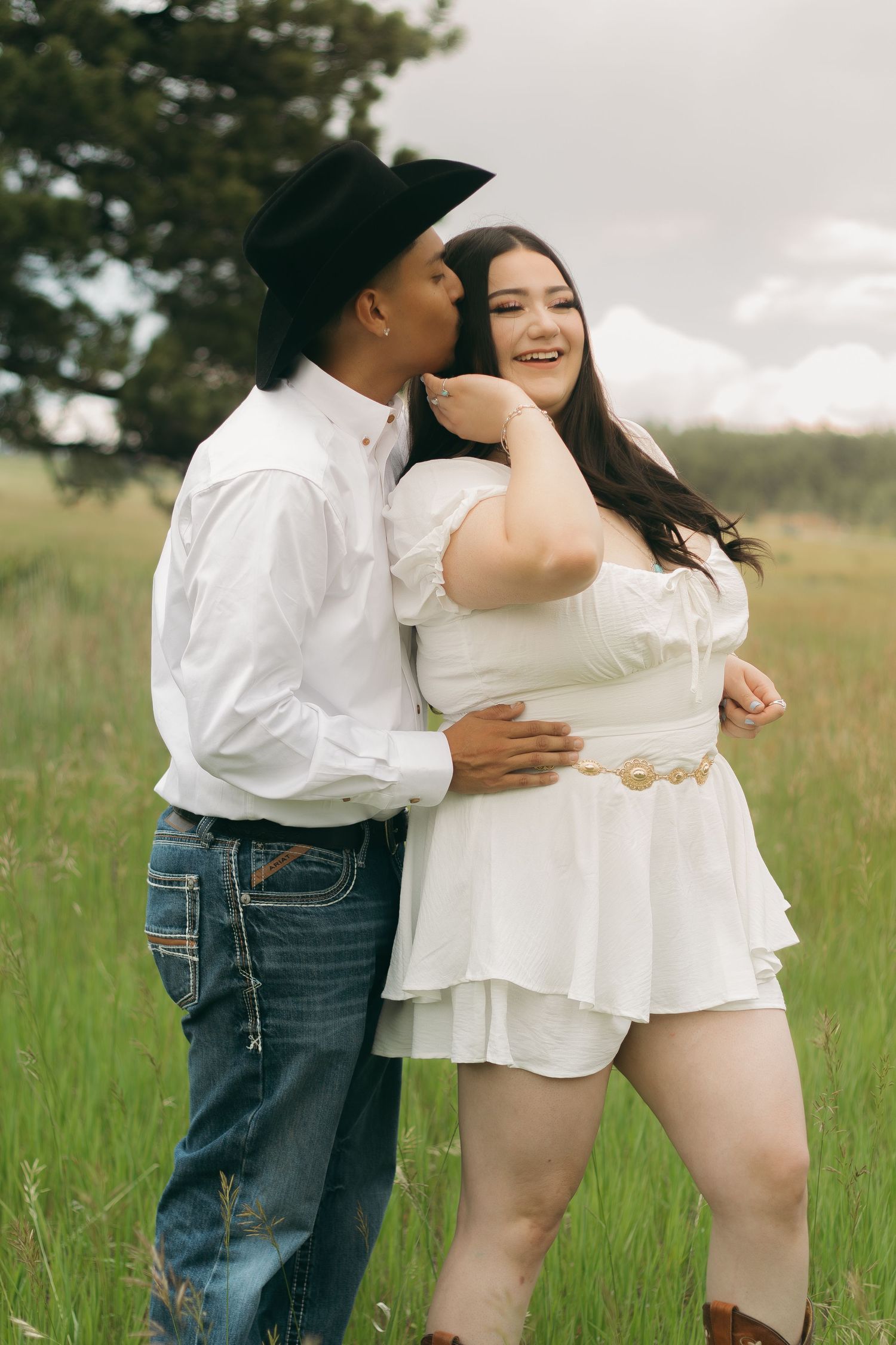 A tender embrace between a couple wearing white outfits in a serene meadow setting.
