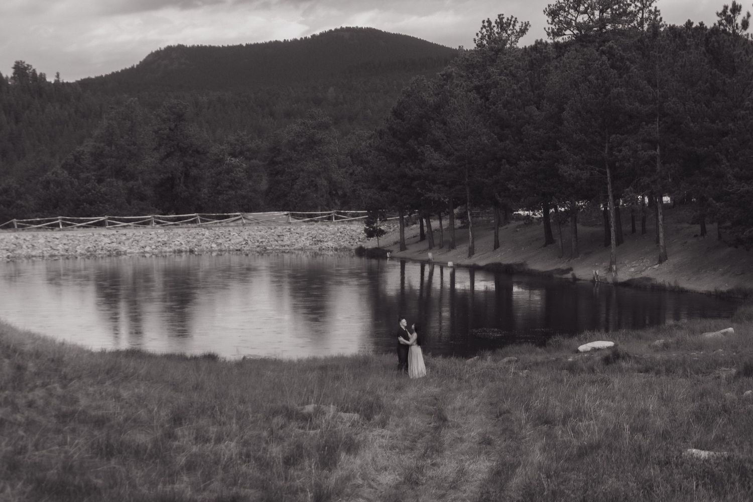 A black and white landscape photo of a couple near a serene lake surrounded by mountains and pine trees.