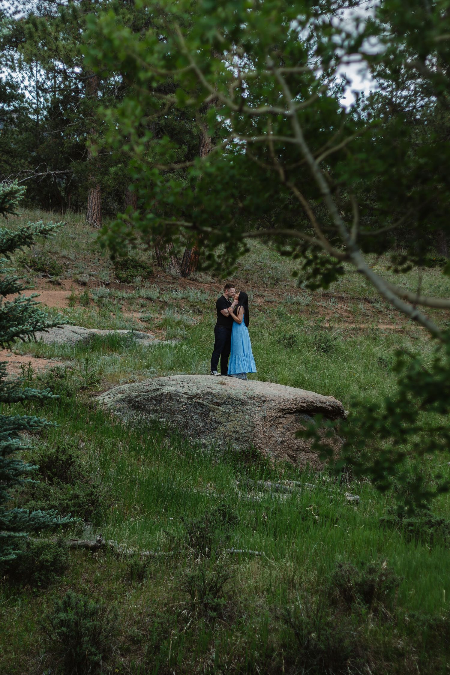 A couple stands together on a large boulder surrounded by evergreen trees and natural scenery.