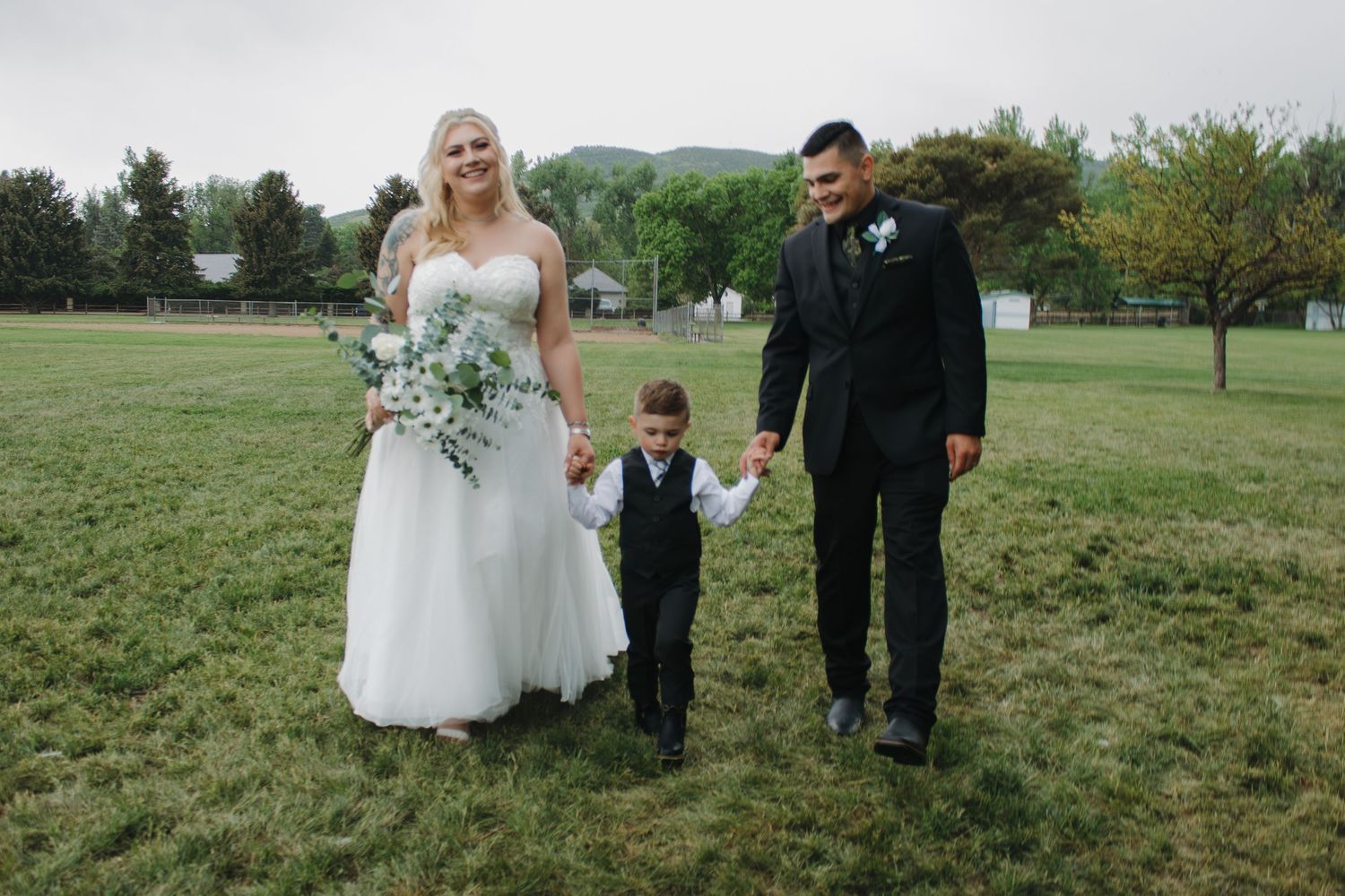 Wedding party members walking together across green field with mountains in background.
