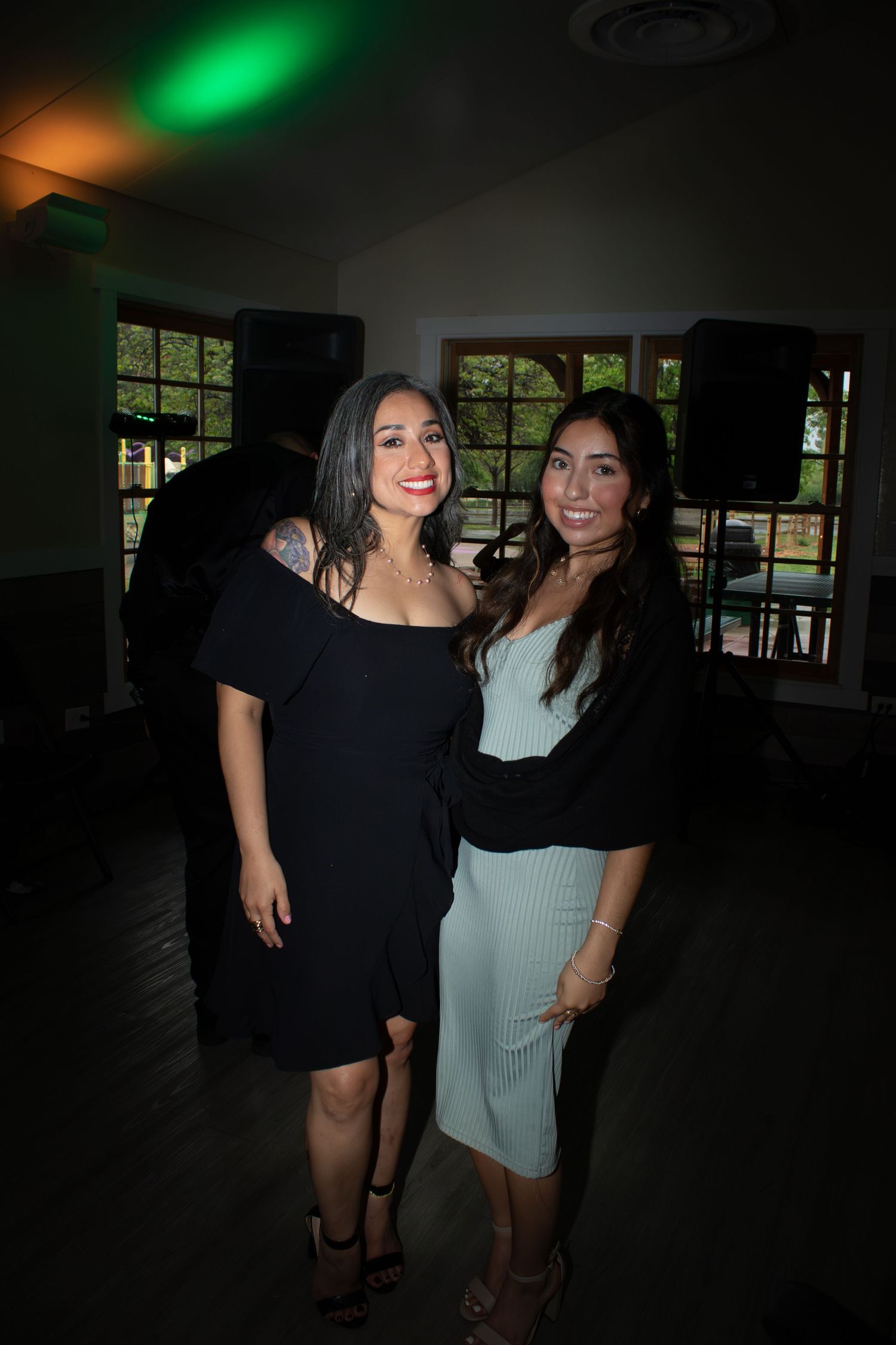 Two women smile together at a dimly lit indoor venue with green accent lighting.