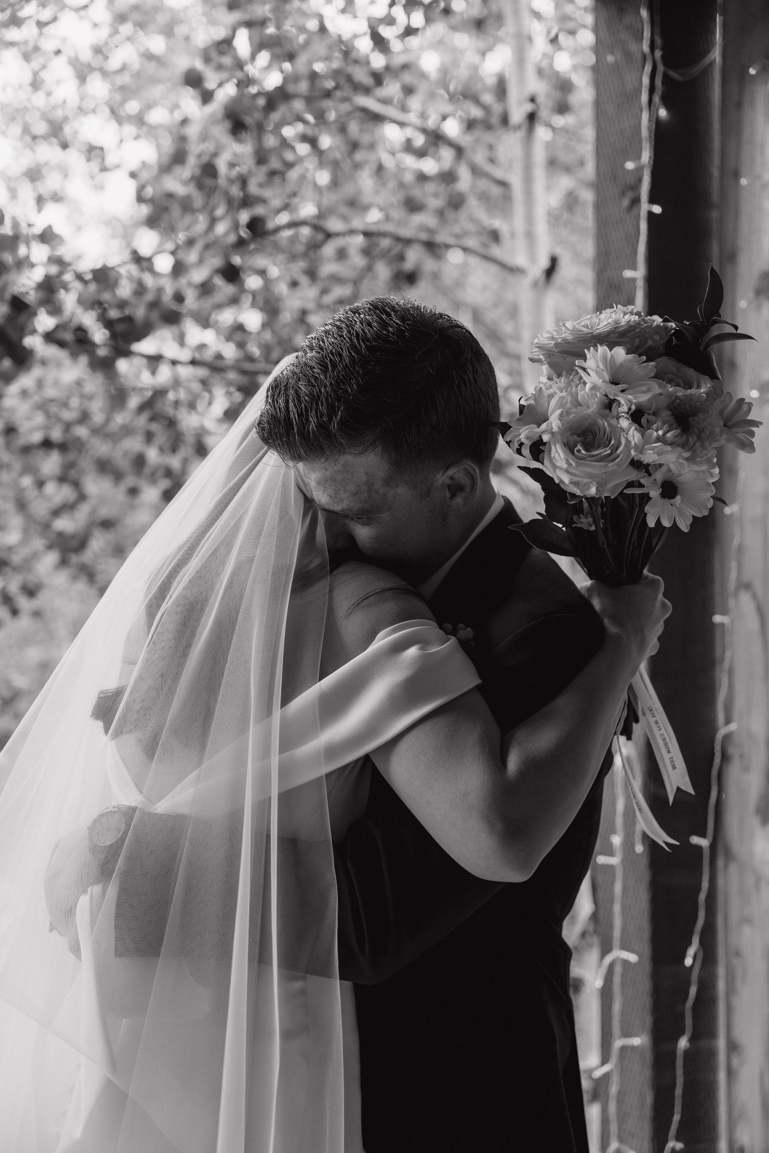 Black and white photo of bride and groom embracing, bride holding bouquet with veil flowing, surrounded by trees.