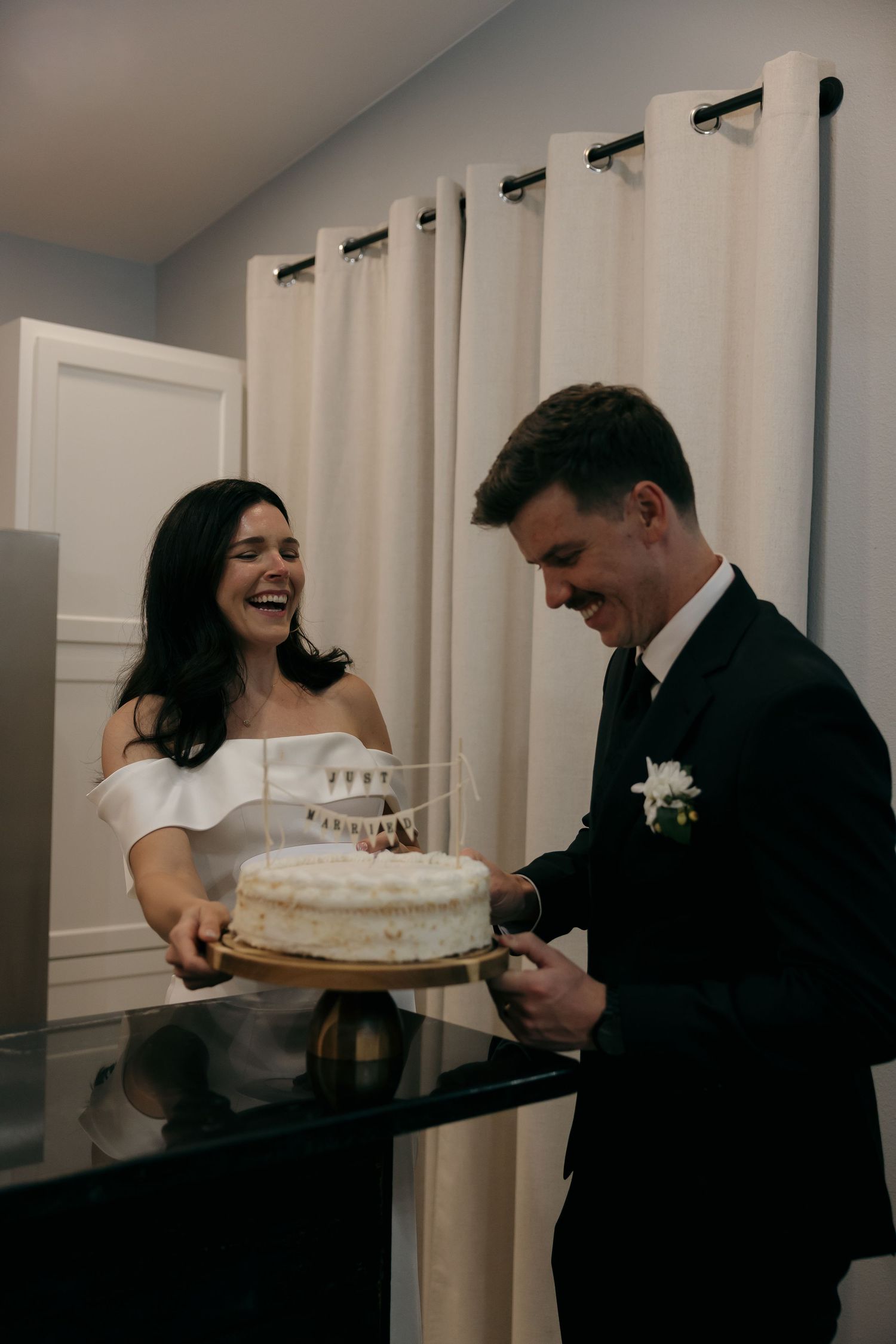 Bride and groom laughing while cutting their white wedding cake indoors at their reception.
