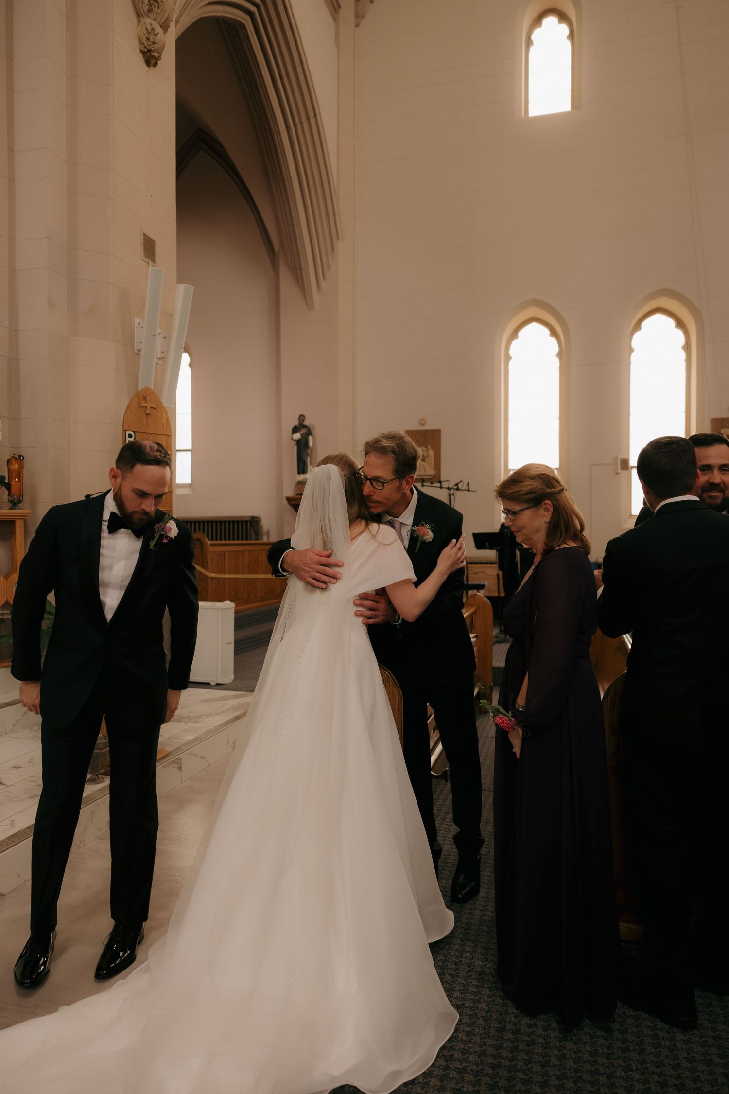 Bride and groom embracing family members inside elegant Catholic church during wedding ceremony.