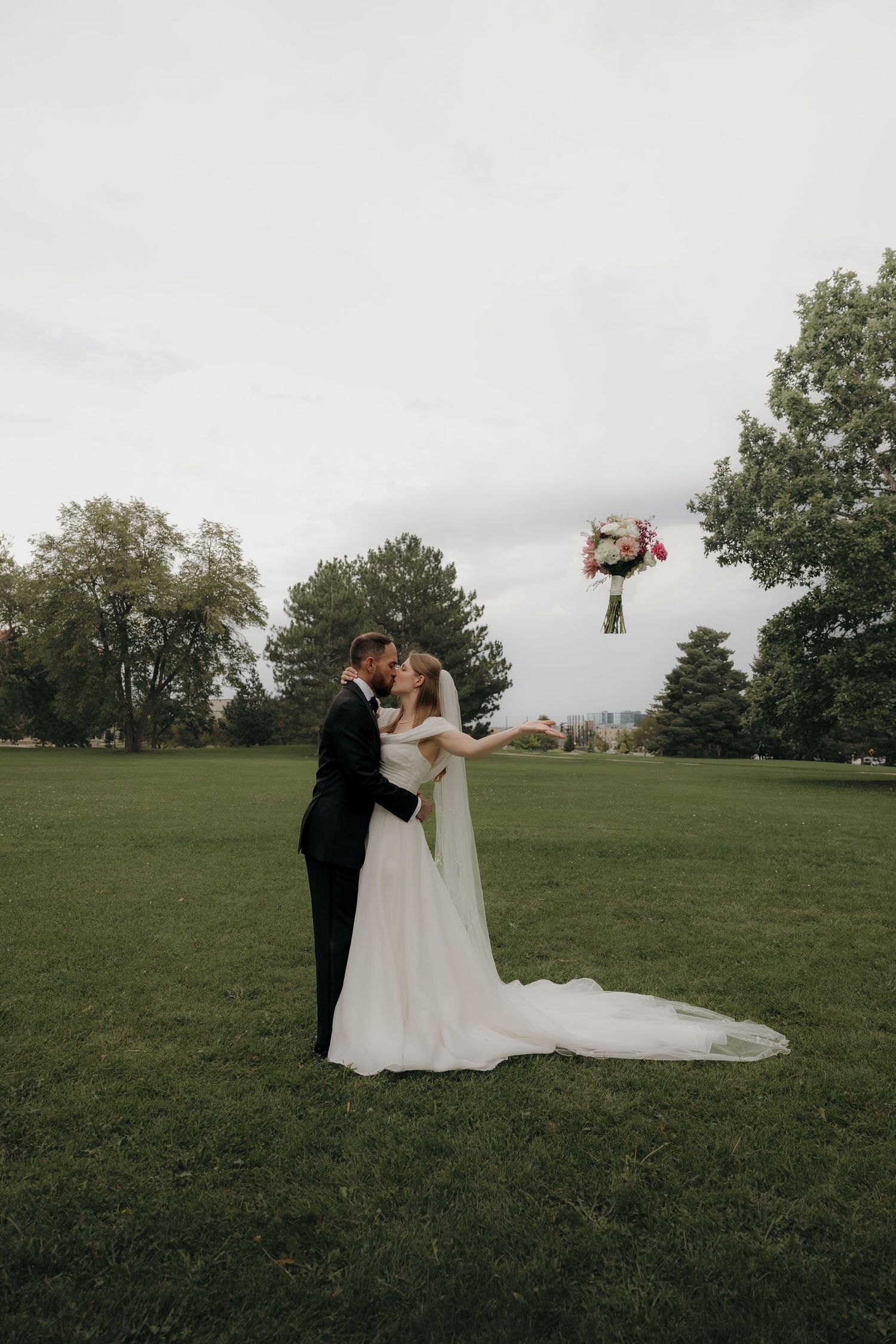 Bride tosses pink and white bouquet while kissing groom in elegant white gown in park setting.