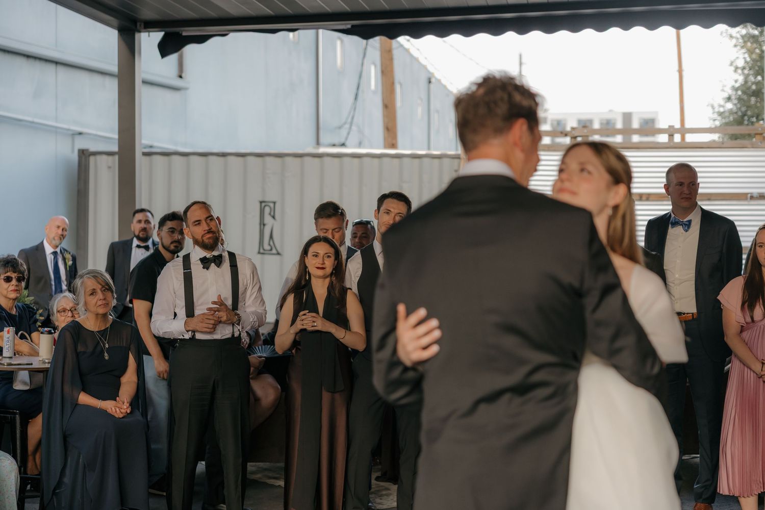Wedding guests watch couple share first dance at outdoor industrial venue reception.