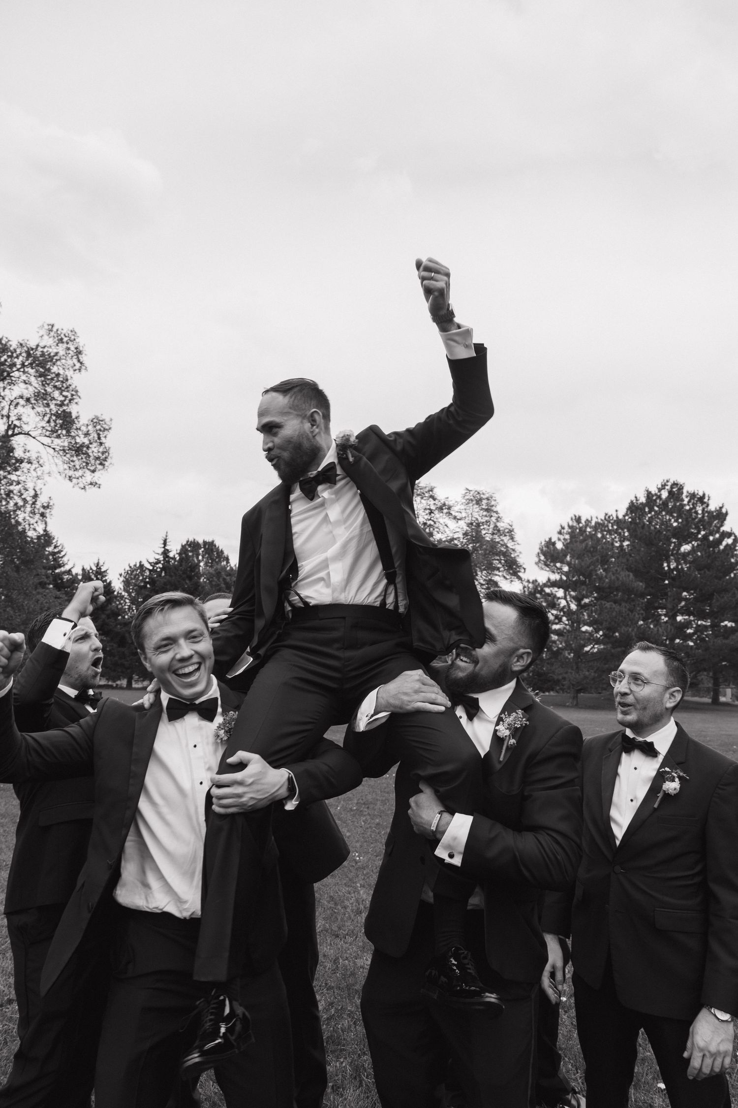 Groomsmen lift celebrating groom in black tuxedo in black and white photo.