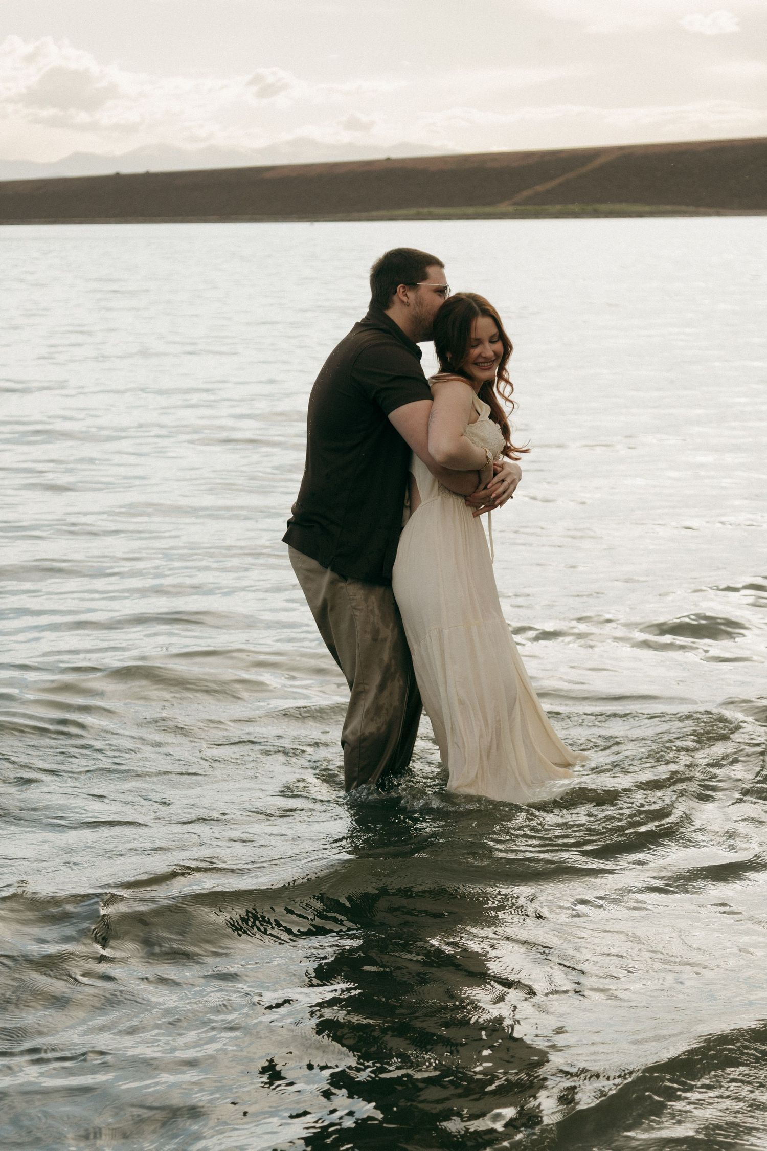 Romantic couple embracing in shallow lake water at sunset, man kissing woman in flowing white dress.