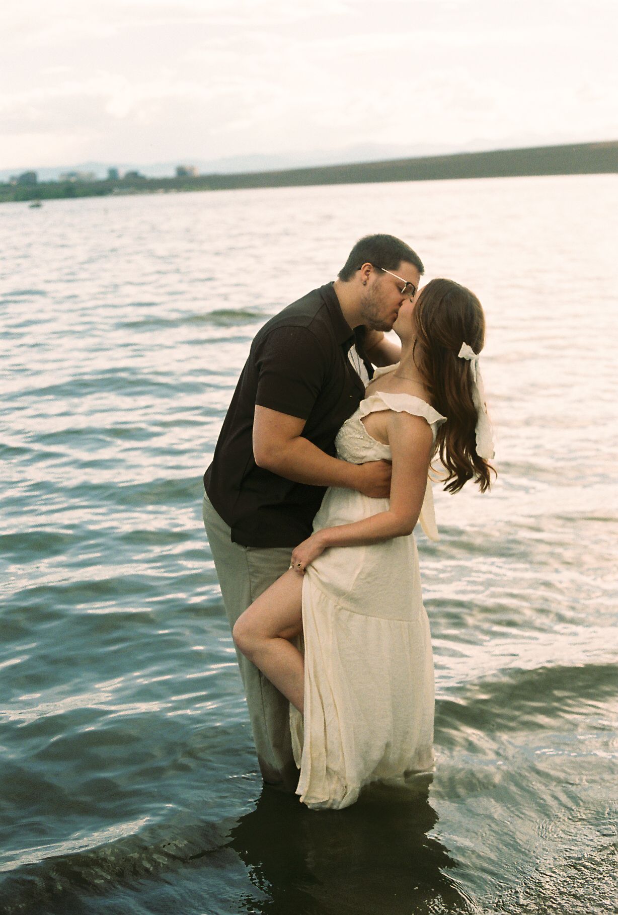 Couple sharing a romantic kiss while standing waist-deep in a lake at sunset, woman in white dress and man in black.