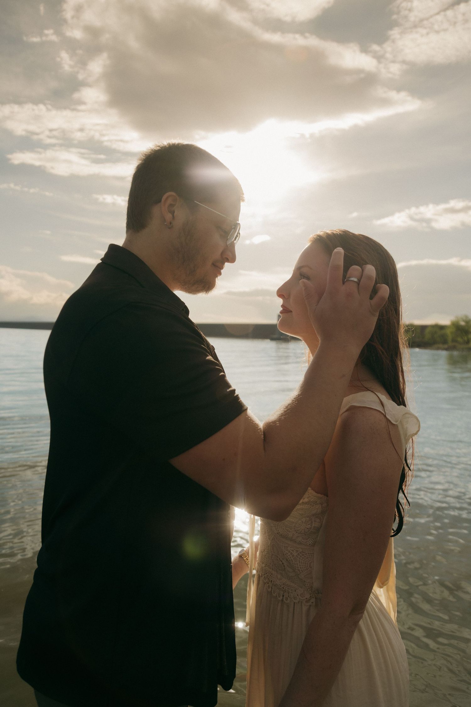 Man tenderly brushing woman's wet hair back by a sunlit lake with dramatic backlit golden hour glow.