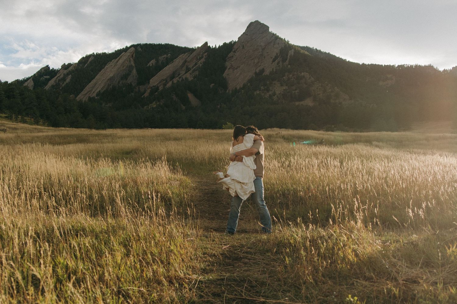 Couple dancing in a sunlit golden field with dramatic rocky flatiron mountains in the background.