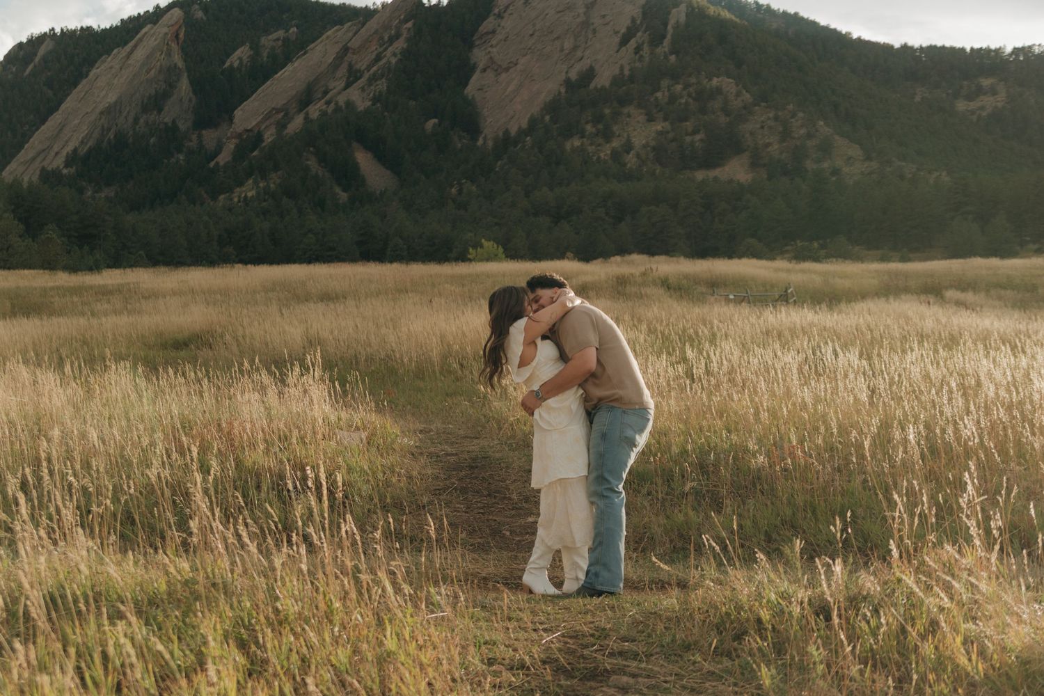 Couple sharing a romantic embrace on a dirt path in a golden meadow with mountain backdrop.