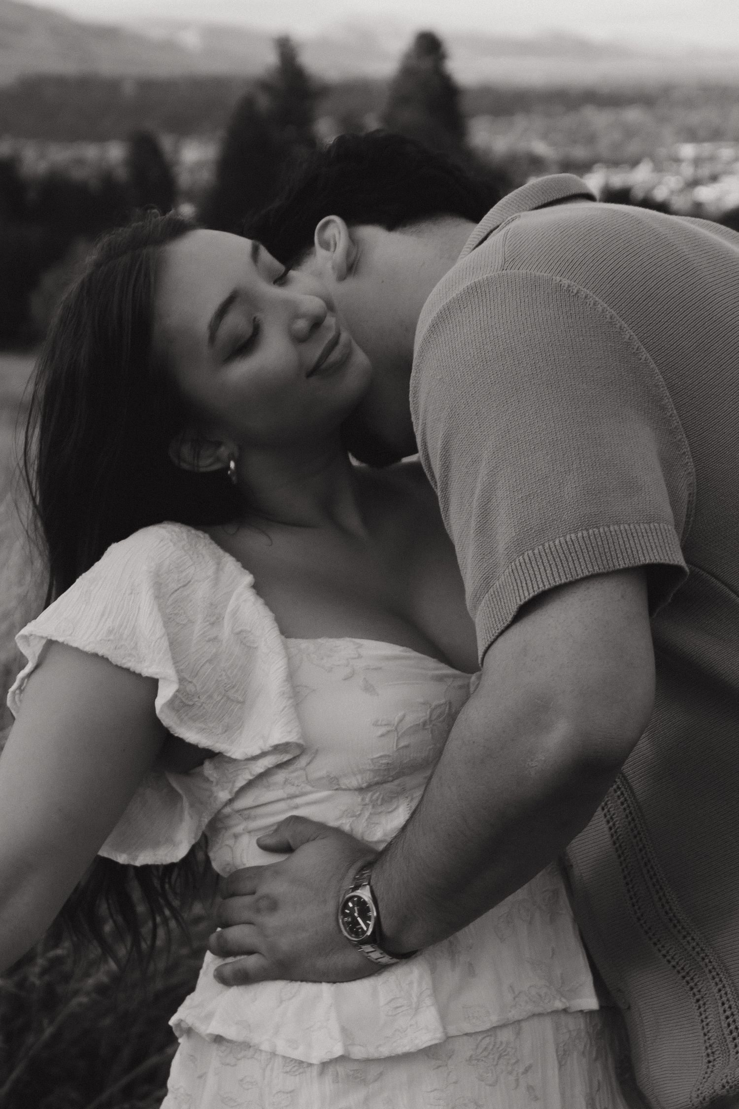 A black and white romantic photo of a couple embracing closely outdoors with a scenic background.