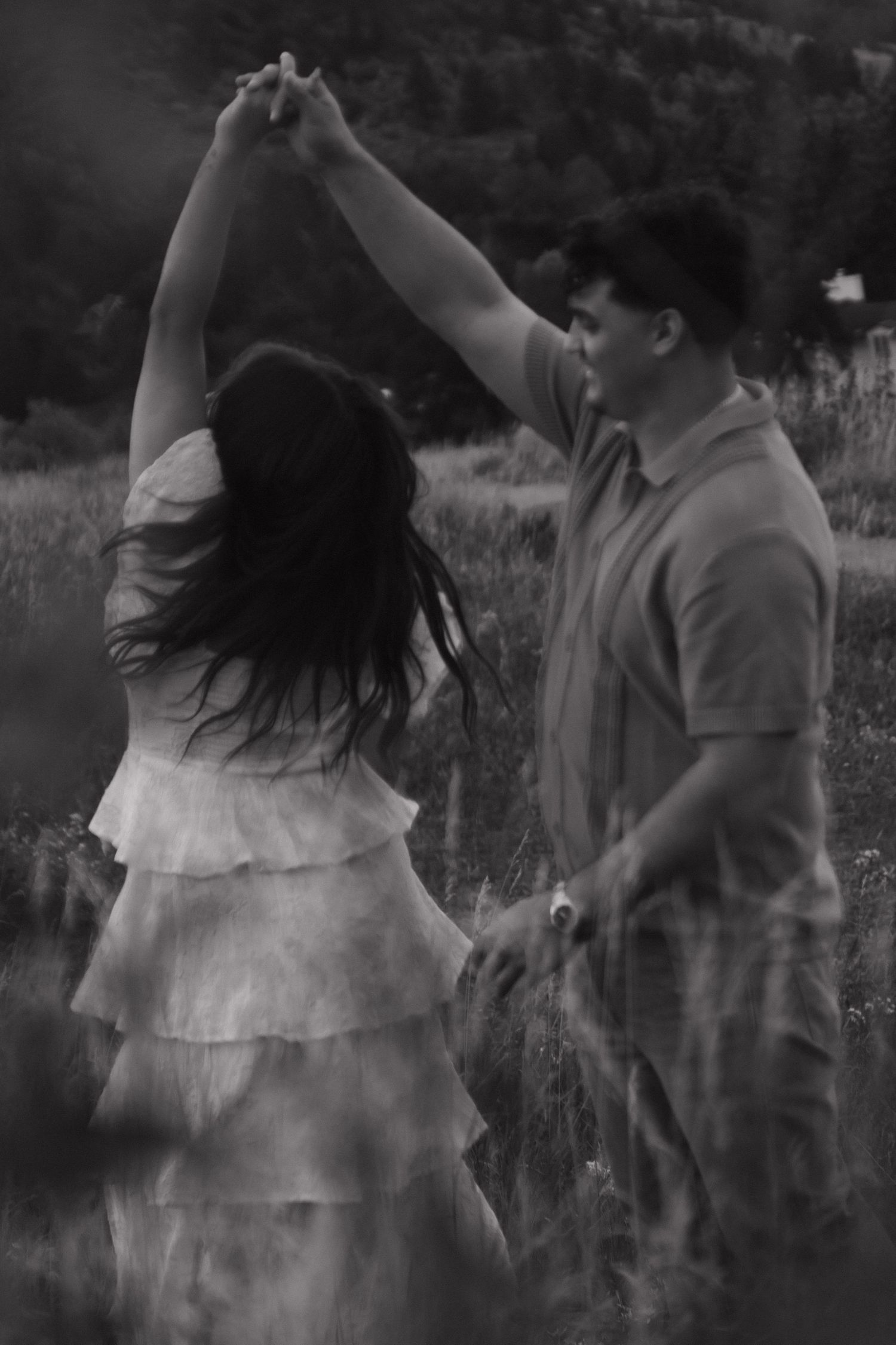 Black and white photo of couple dancing in a meadow, woman twirling in white dress.