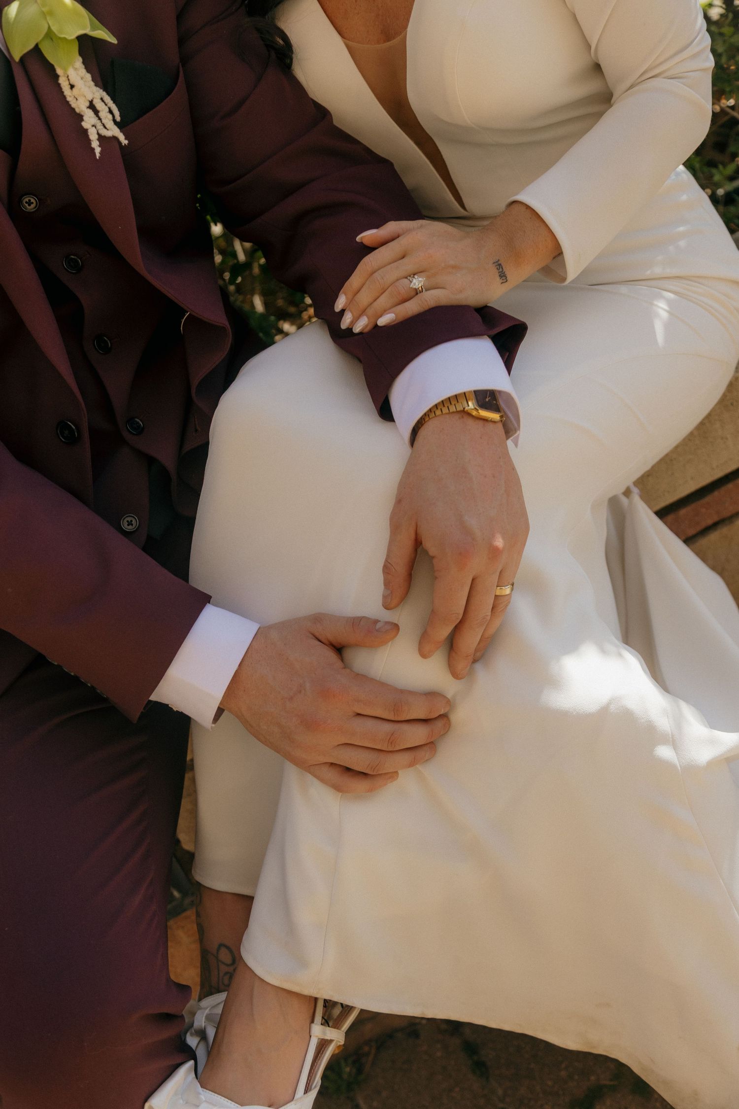 Close-up of bride and groom hands showing wedding rings, bride in white dress, groom in burgundy suit.