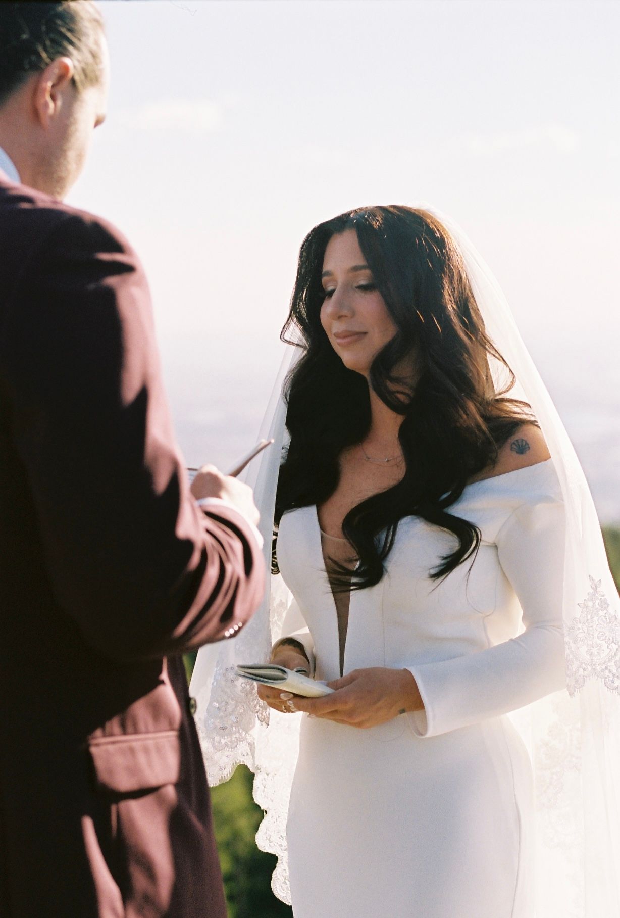 Bride in white off-shoulder gown reads vows from card during outdoor wedding ceremony at sunset.