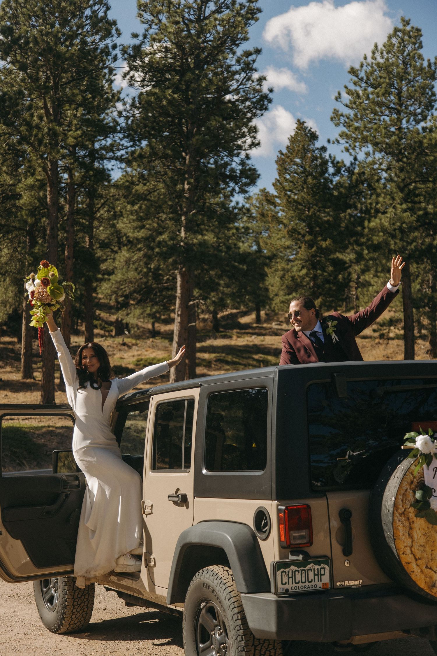 Joyful bride and groom celebrate atop a Jeep Wrangler in a pine forest setting.