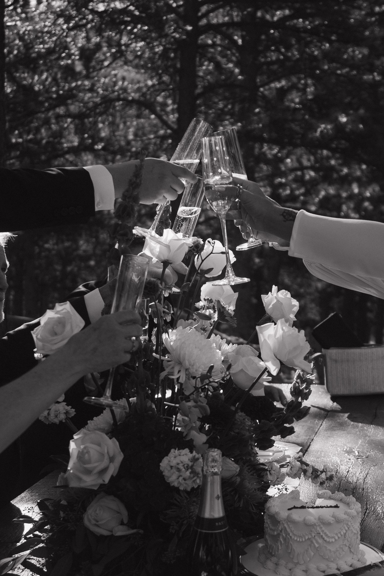 Black and white photo of wedding guests toasting champagne glasses over floral centerpiece and white cake.