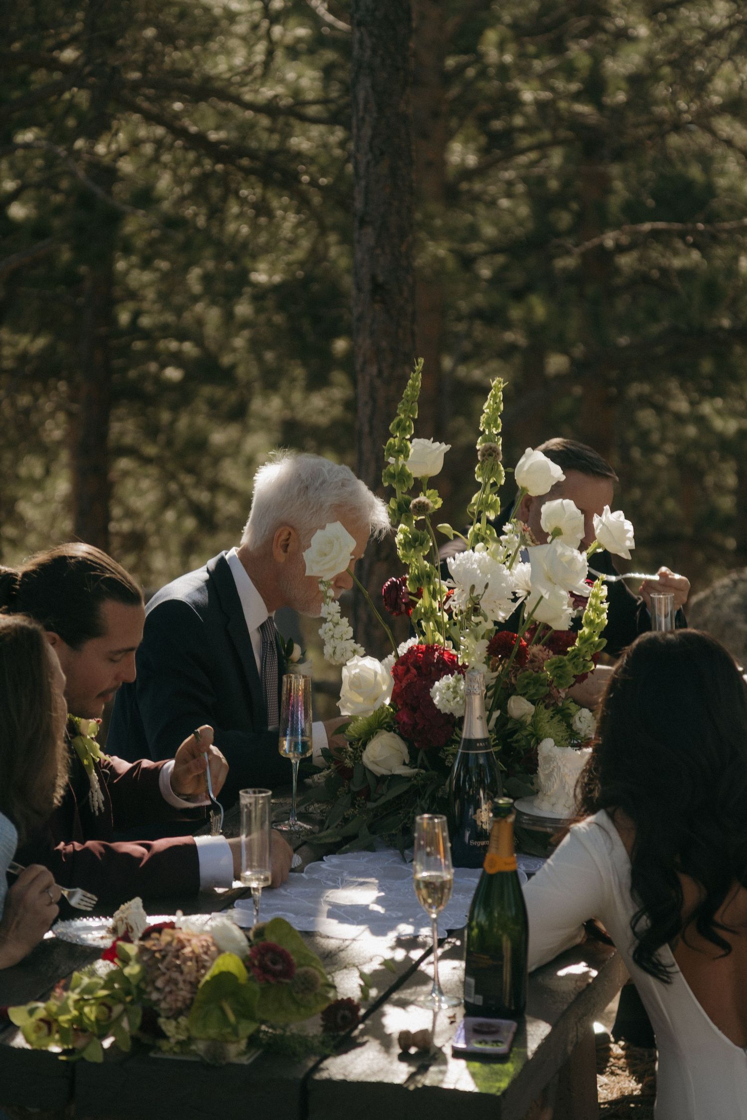Wedding guests dine at an elegant outdoor table adorned with white roses and greenery in a forest setting.