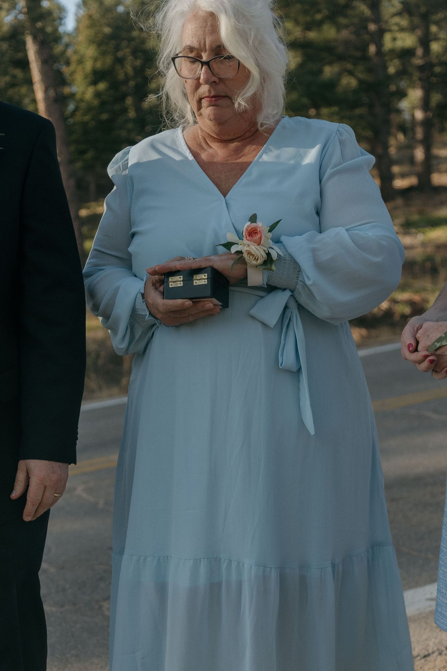 Older woman in light blue dress holding ring box with floral corsage at outdoor wedding ceremony.