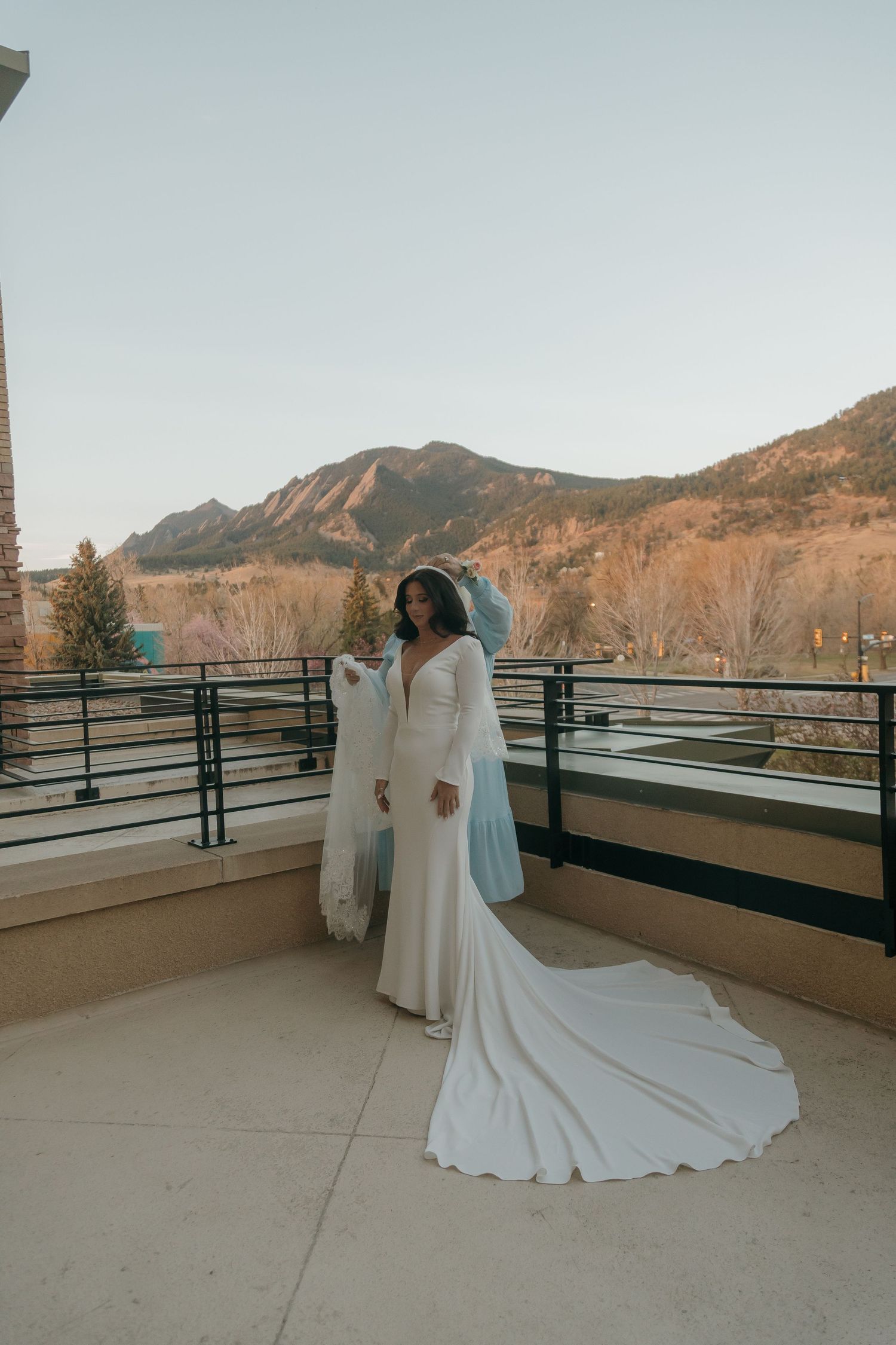 Bride in a white wedding gown with a long train poses on a balcony with mountain views.