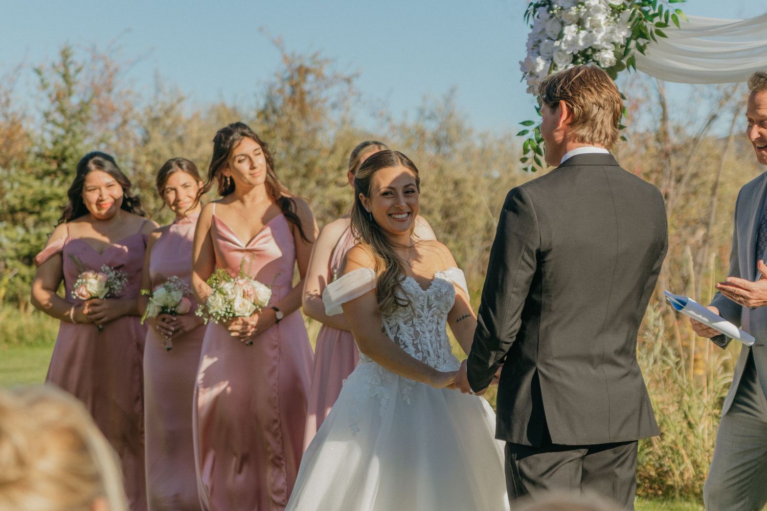 A wedding party in mauve bridesmaid dresses poses together outdoors on a sunny autumn day.