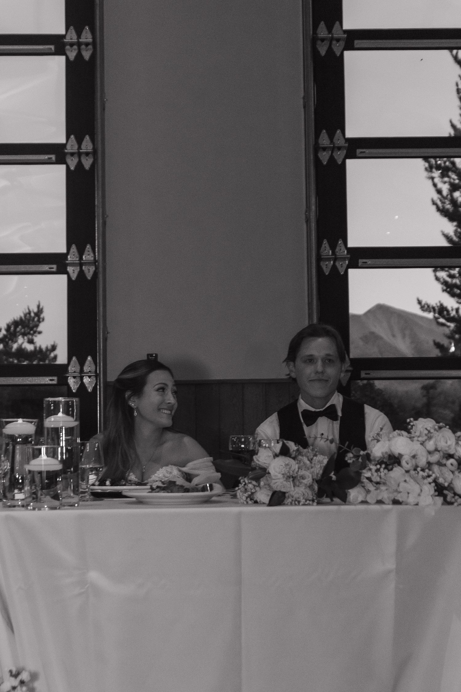 Wedding reception head table setup with centerpieces and place settings against dark backdrop.