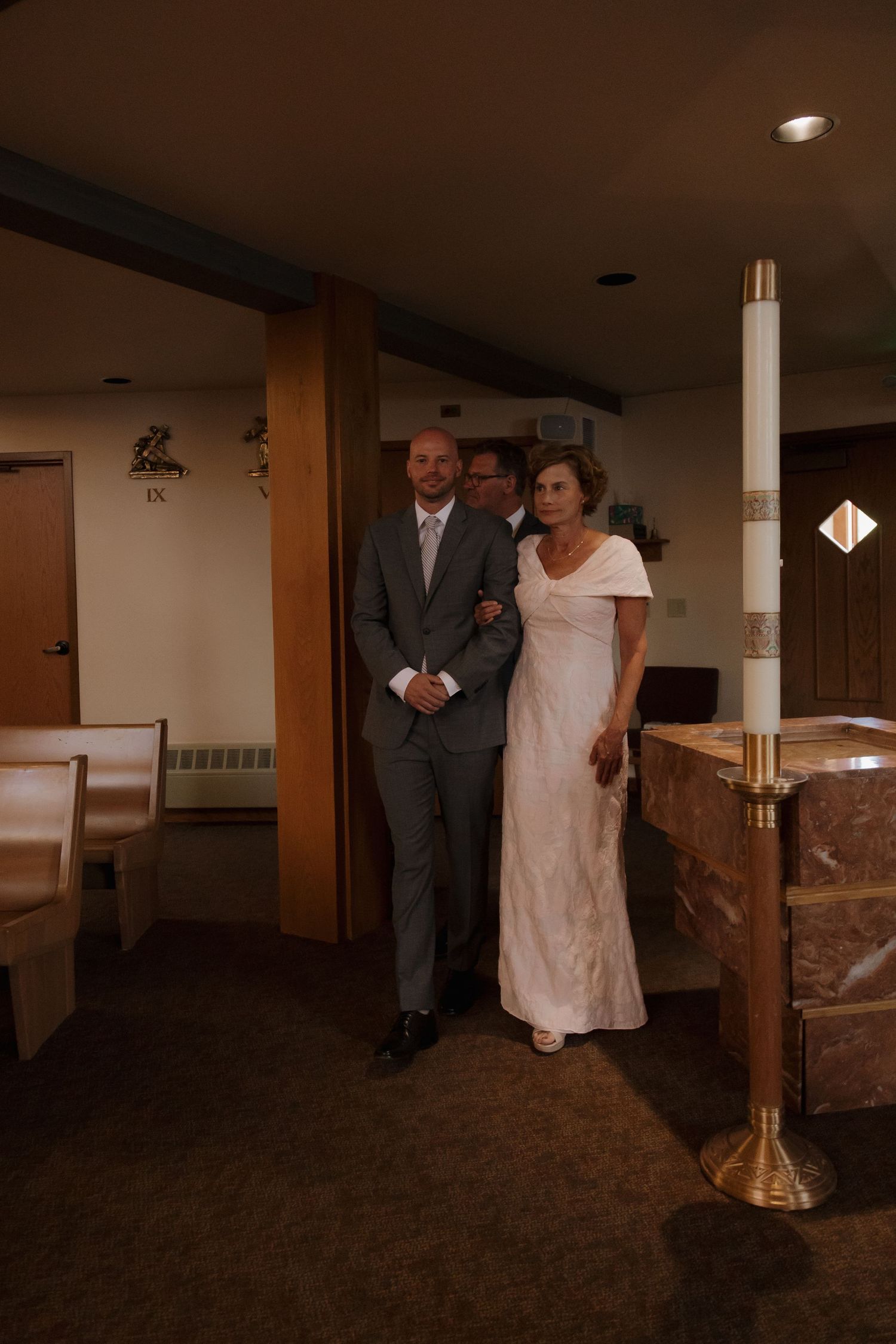 A groom in a gray suit escorts a woman in a white dress down a church aisle during a wedding ceremony.