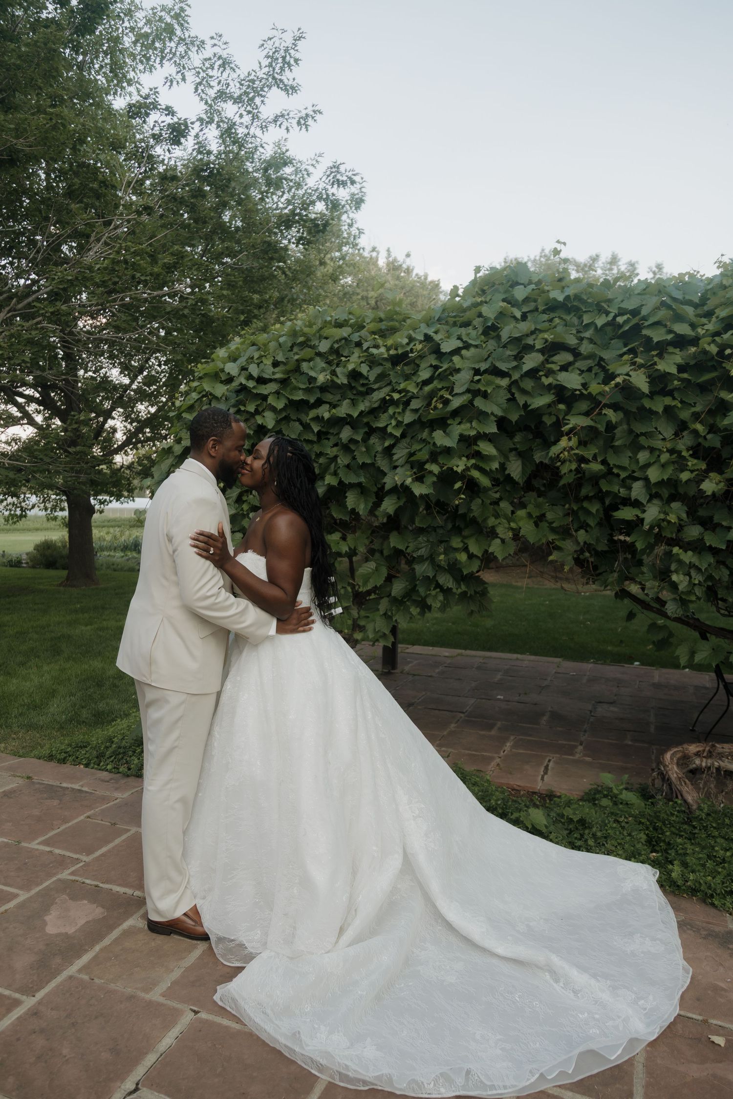 Black bride and groom share a kiss while embracing near lush green vines and trees on their wedding day.