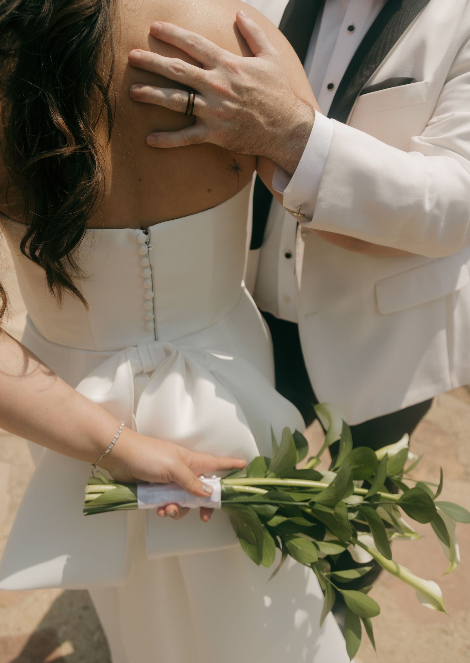 Close-up of bride and groom holding green bouquet, groom's hand resting on bride's bare shoulder.