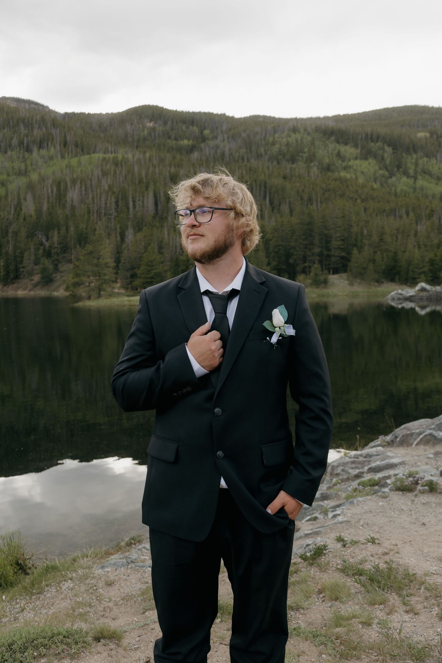 Groom in black suit with boutonniere adjusting tie beside a reflective mountain lake with forested hills.