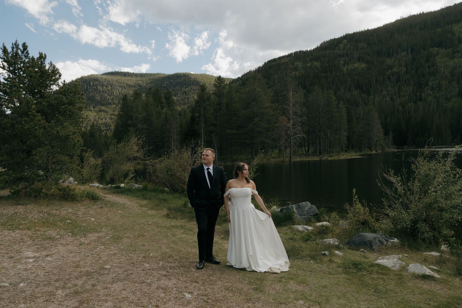 Bride and groom walk hand-in-hand along a mountain trail beside a scenic alpine lake.
