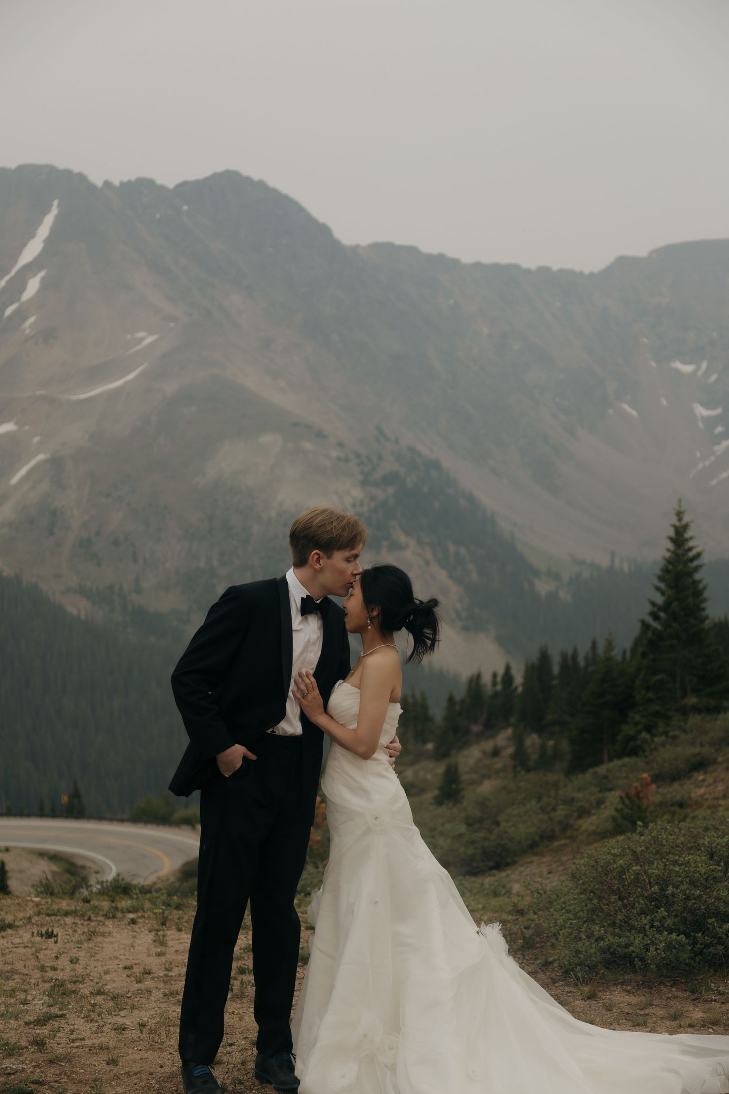 Bride and groom share a romantic kiss with misty mountain peaks and evergreen trees in the background.