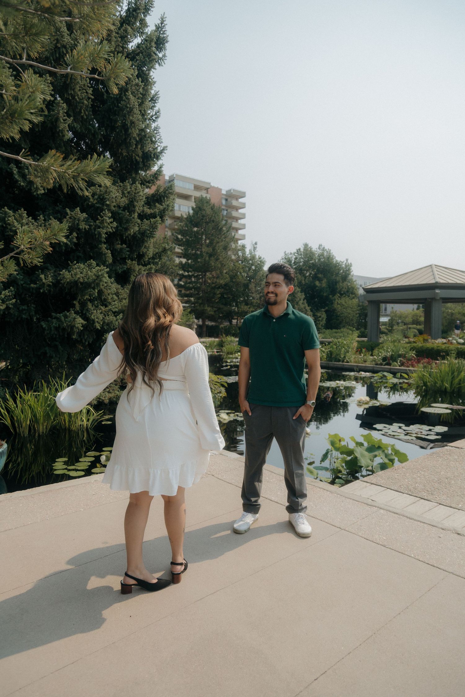 Couple strolling by a serene garden pond, woman in white dress walking toward smiling man in green polo shirt.