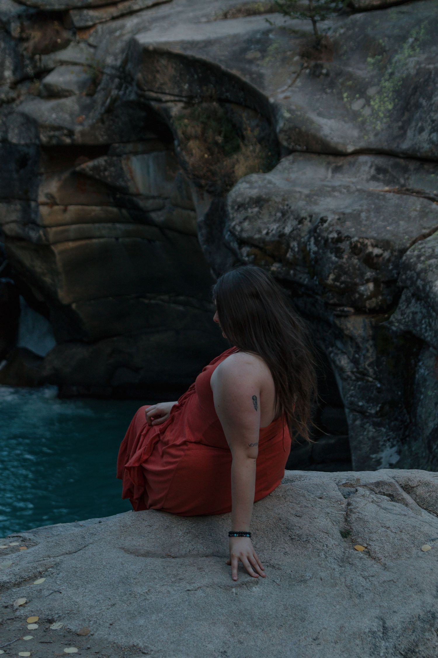A person in a red dress lies on a rocky ledge overlooking turquoise waters in a dramatic natural setting.