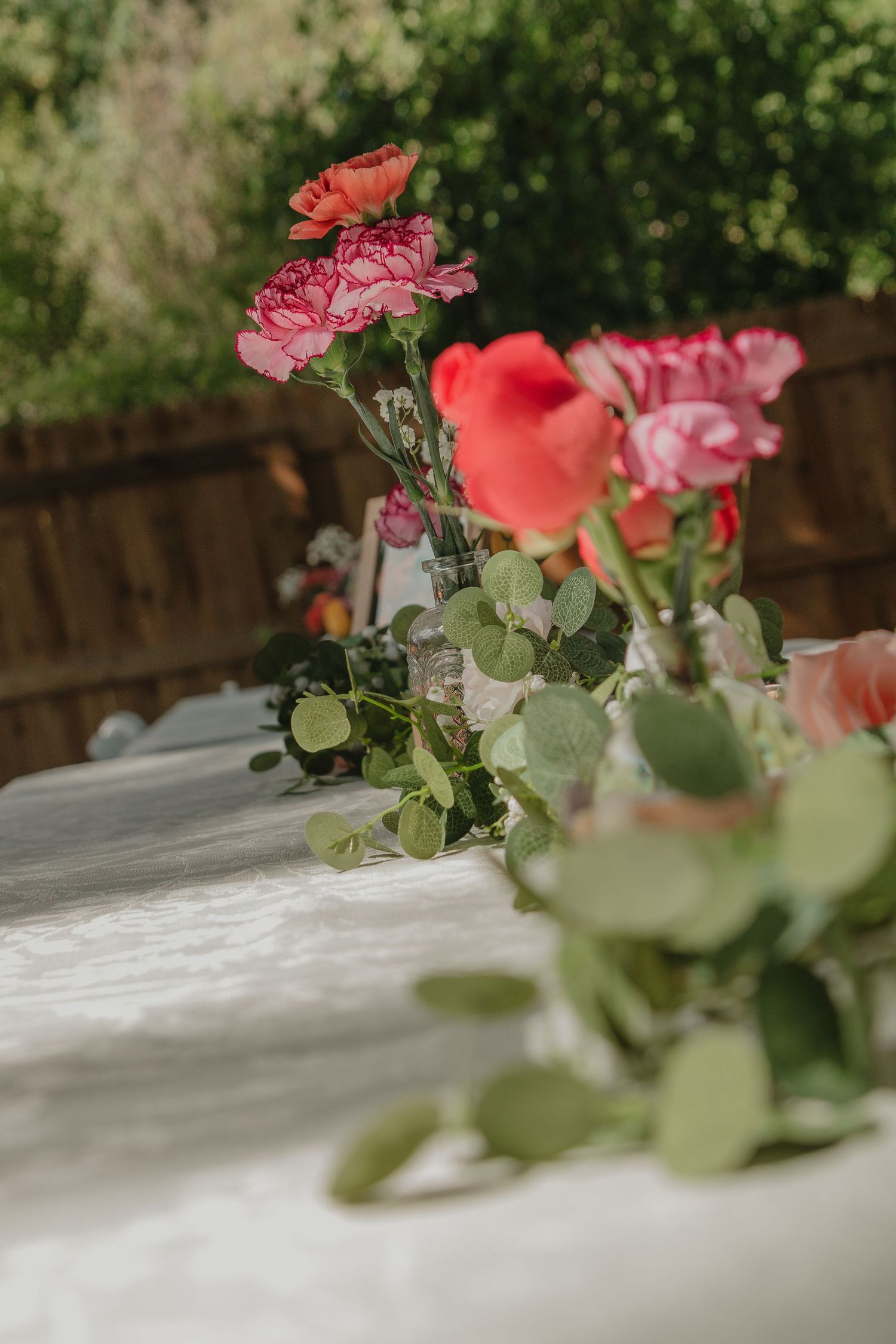 Pink and coral carnation flowers arranged on a white tablecloth with soft green foliage.